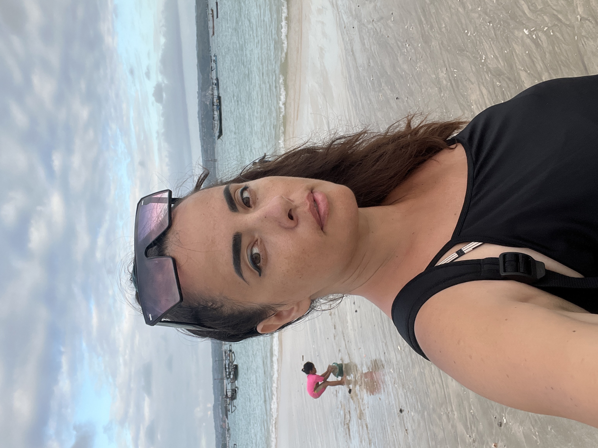 In Kuta, Indonesia, a woman wearing sunglasses on her head and a black tank top stands on a wet sandy beach. The ocean is behind her with several boats in the distance. A young child in a pink swimsuit is playing in the shallow water to the left of the woman. The sky is cloudy and the lighting suggests it is late afternoon or early evening.