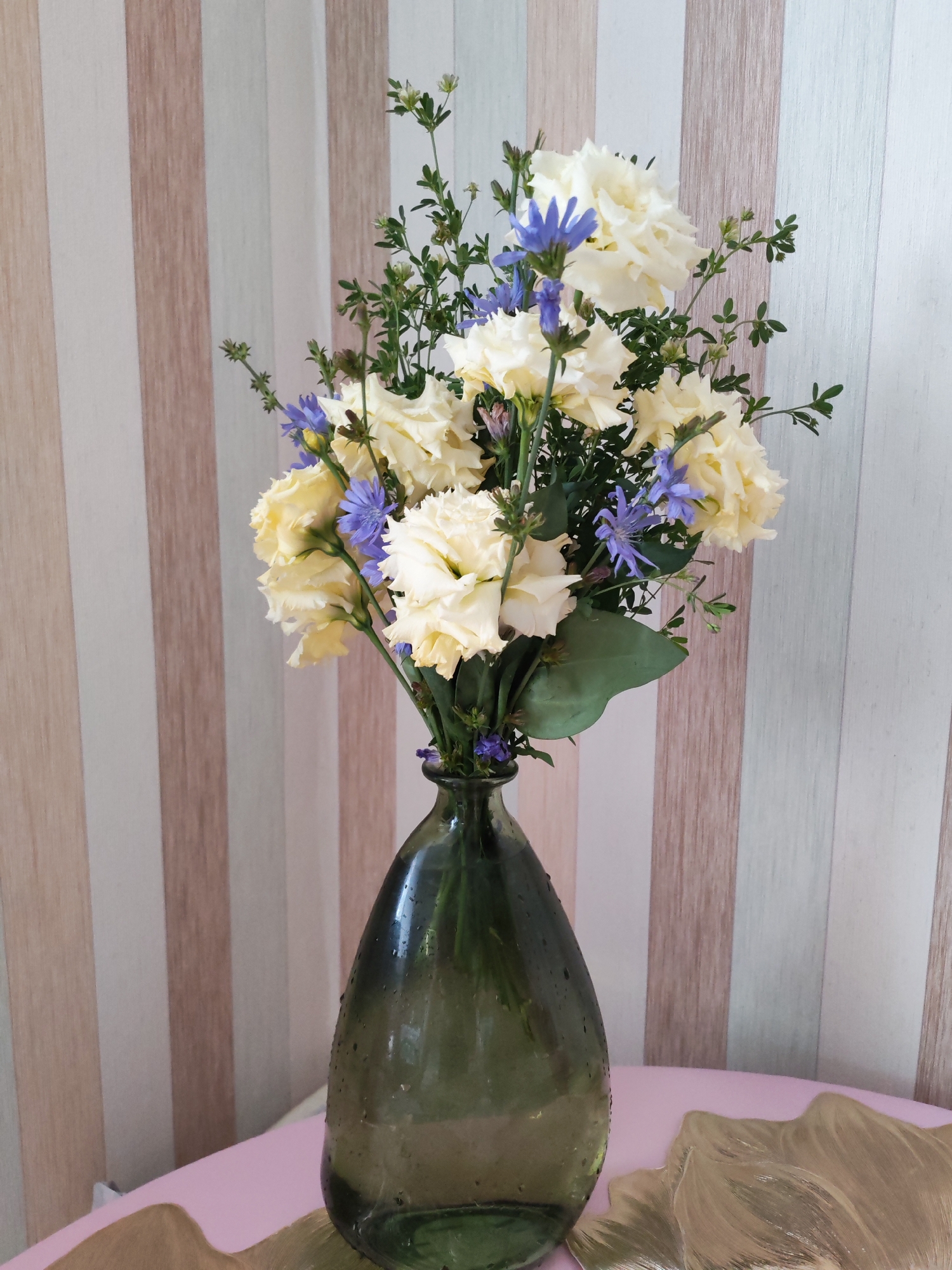 A bouquet of light yellow flowers and delicate blue wildflowers is arranged in a dark green glass vase. The vase is on a light pink surface, and behind it is a wall with vertical stripes in shades of beige, brown, and off-white. The image was taken in Izhevsk, Russia.