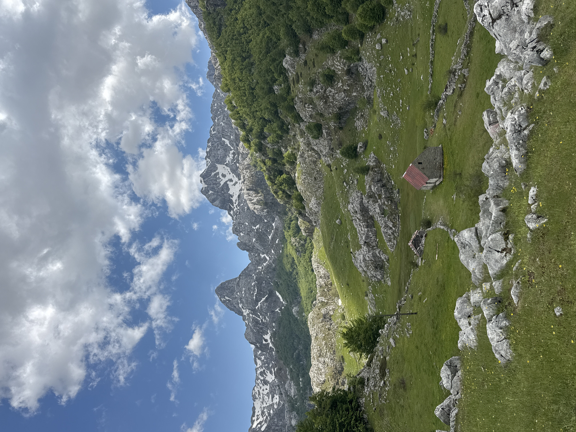 The image depicts a mountainous landscape in Brskut, Montenegro. Rocky, snow-dusted mountains rise on the left under a blue sky with scattered clouds. The right side features a grassy hillside with exposed rocks. A small stone building with a red roof is nestled into the landscape. A low stone wall runs along the hillside. Trees are present among the rocks.