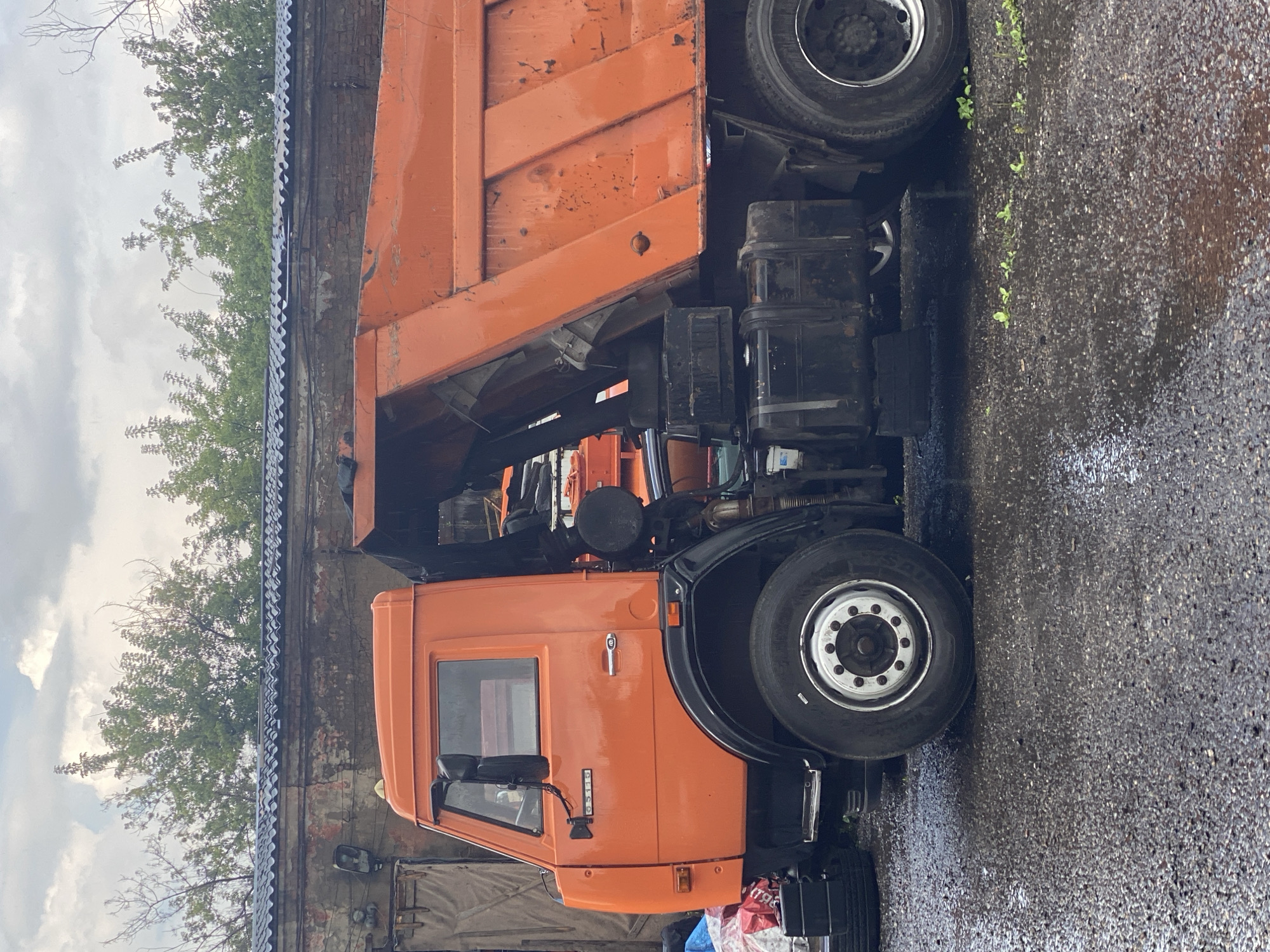 The image shows an orange dump truck parked on a wet, gravelly surface, likely a service area or lot. The truck's bed is raised, indicating it may have recently dumped material or is being serviced. The background features a brick building with a corrugated metal roof and lush green trees. The sky is cloudy, suggesting overcast weather. The truck appears to be in a used condition, with some visible wear and tear on its paint. The overall scene suggests an industrial or utility setting within Novokuznetsk, Russia.