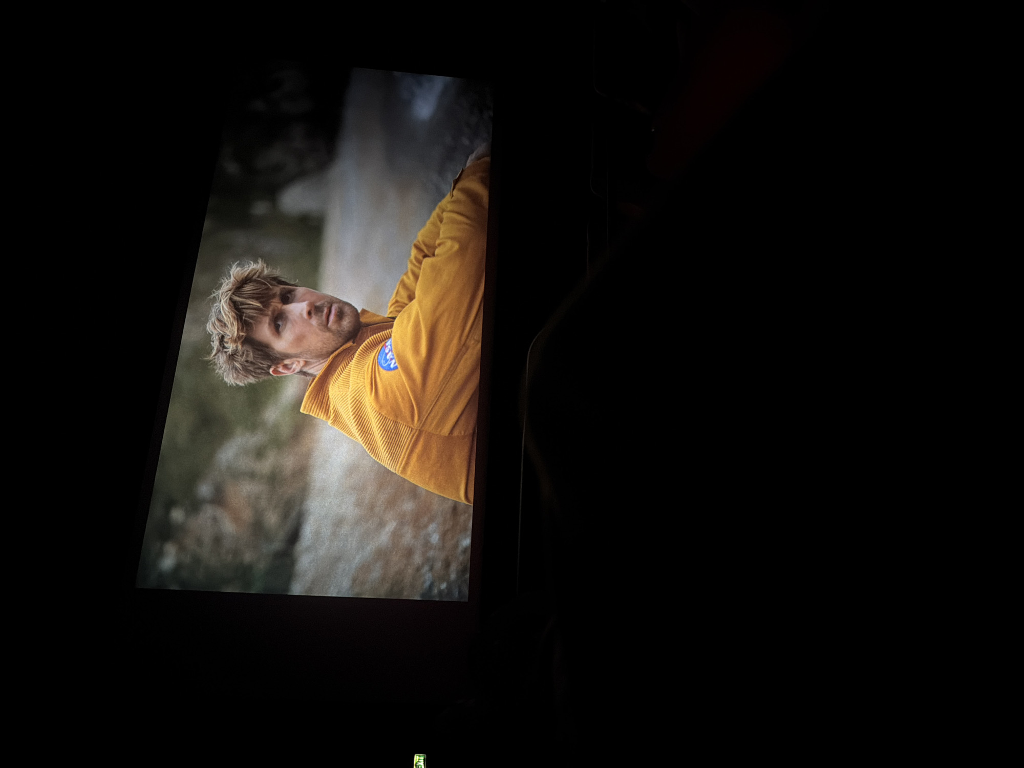 The image shows a movie screen in a dark room. A man is featured on the screen, likely an actor in the movie. He has blond hair and is wearing a yellow corduroy jacket with a blue patch. The background behind him appears to be a nature scene with rock formations and possibly vegetation. The room is largely dark, with only the movie screen providing illumination. The location in Sofia, Bulgaria cannot be determined from the visual information provided.