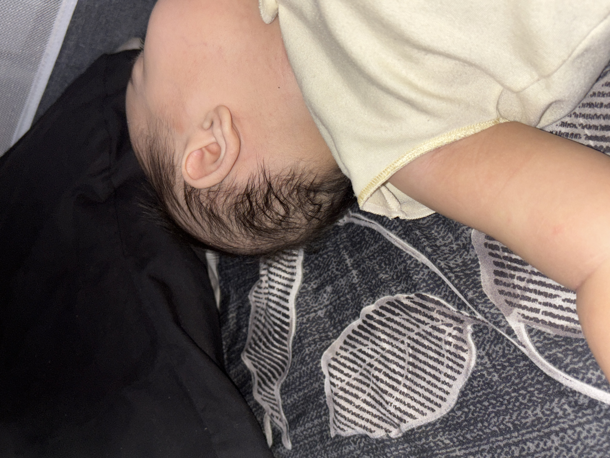 A close-up, eye-level shot shows the head and upper body of a baby sleeping on a patterned blanket. The baby's head is turned to the right, with the ear clearly visible. The baby has dark hair, and is wearing a light yellow onesie. The blanket is dark gray with a white and black striped pattern. A dark fabric, possibly a pillow or blanket, is visible in the upper left corner of the image. The scene appears to be indoors, in a residential setting. This image is not taken in the city of West Jakarta, Indonesia.