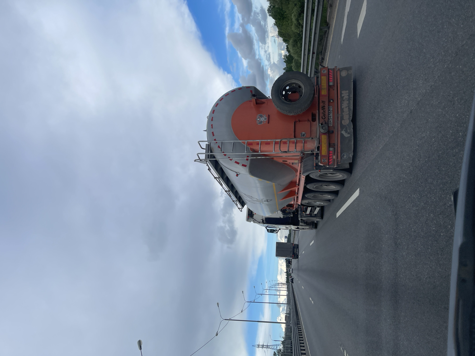 The image shows a grey and orange GuteWolf tanker truck traveling on a highway near Бавыкино, Russia. The truck has a spare tire mounted on the back and ladder access to the top of the tanker. Multiple trucks and cars are driving in the same direction on the highway. Street lights line the side of the road and a forested area is visible on the other side of a barrier. The sky is partly cloudy.