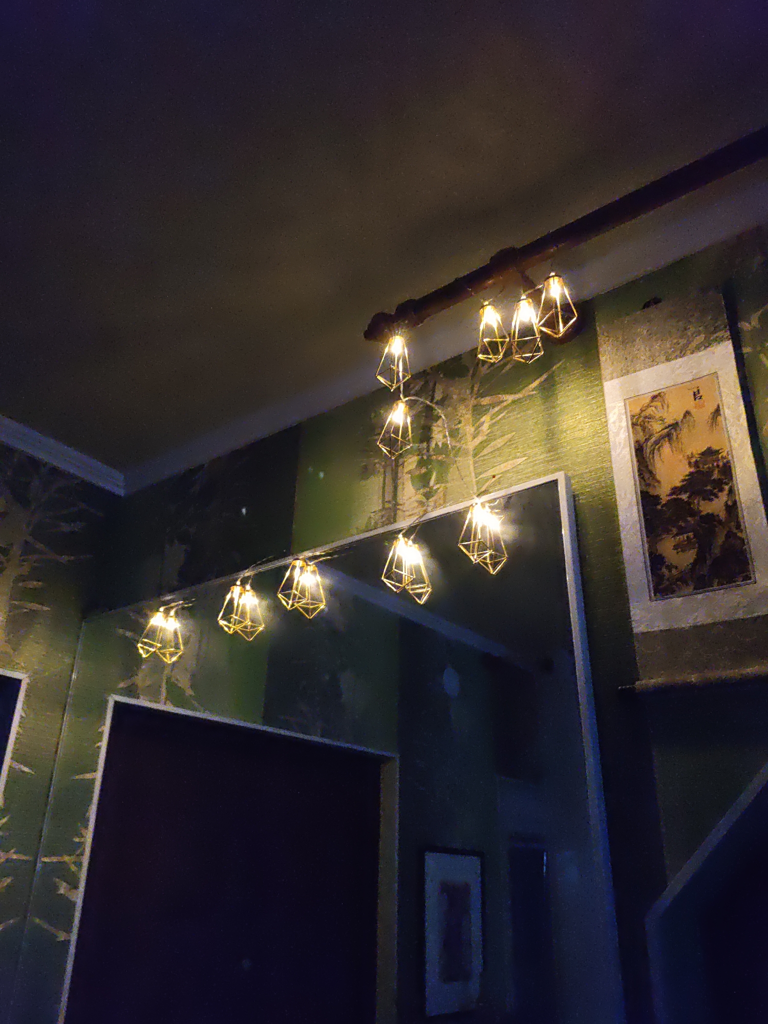 This is a low-angle, indoor shot of a room, possibly a bar or restaurant, with a dark, textured ceiling and green patterned wallpaper. A series of decorative pendant lights, shaped like geometric cages with visible bulbs, hang from a dark wooden beam across the upper portion of the frame. The lights are illuminated with a warm, yellow glow. Below the lights, a large mirror reflects the same light fixtures and the green wallpaper, creating a sense of depth and repeating patterns. To the right of the mirror, a framed artwork with a landscape scene, rendered in a traditional East Asian style, hangs on the wall. The artwork depicts mountains, trees, and what appears to be a building. The overall atmosphere is dimly lit and suggests a stylized interior design. The image does not provide any direct information about the city of Rostov-on-Don, Russia.