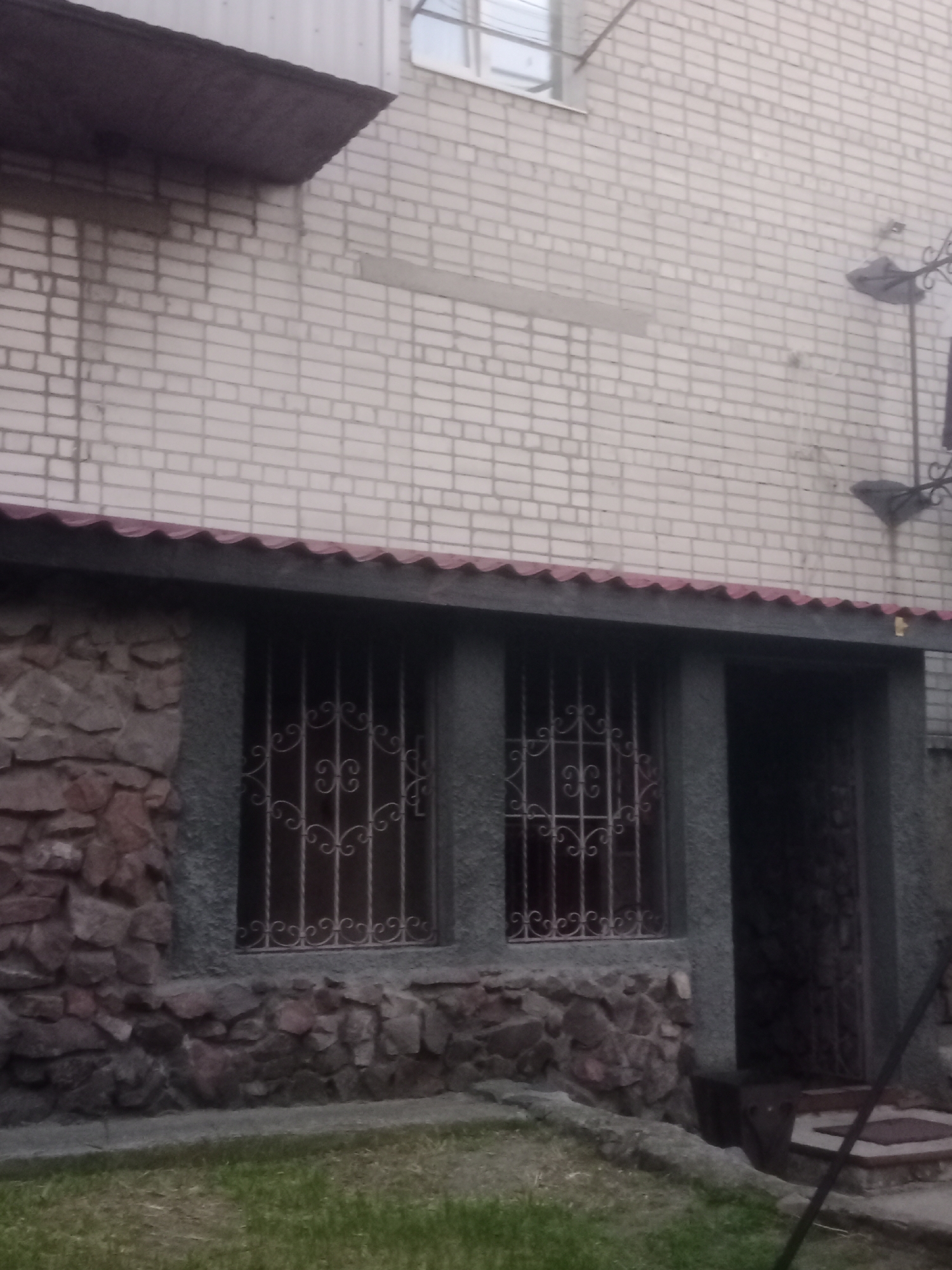 The image shows the exterior of a building, likely in the city of Horishni Plavni, Ukraine. The building has a light-colored brick upper facade and a lower section constructed with rough-hewn stones and finished with a dark grey textured material. Two windows on the lower level are protected by decorative wrought iron bars. Adjacent to the windows is an entrance with a dark door, also featuring decorative metalwork. A dark, wavy roofline extends over the lower section of the building. The ground in front of the building is covered with grass. The lighting suggests it is daytime, possibly overcast.