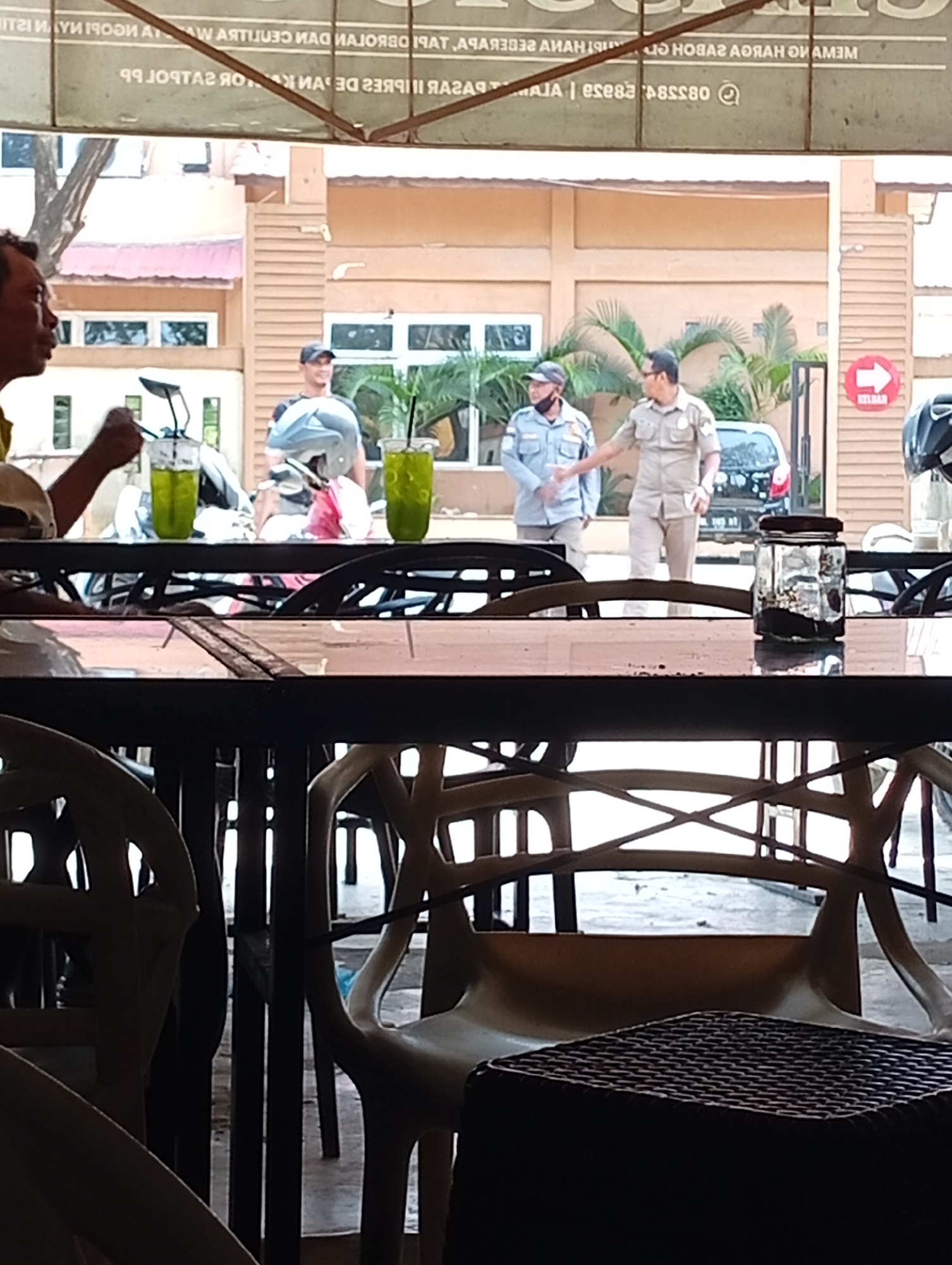 The image depicts an outdoor scene at a roadside cafe or restaurant in Lhokseumawe, Indonesia. In the foreground, a table is set with two glasses of green beverage and a small jar with a dark substance. Several chairs are visible, some empty and some occupied by patrons partially obscured by the table and foreground elements. In the middle ground, two individuals in what appear to be uniforms are engaged in conversation, with one gesturing. Behind them, a motorcycle and a black car are parked. The background shows a building with windows and a red directional sign pointing to the right. The overall atmosphere suggests a casual daytime setting.