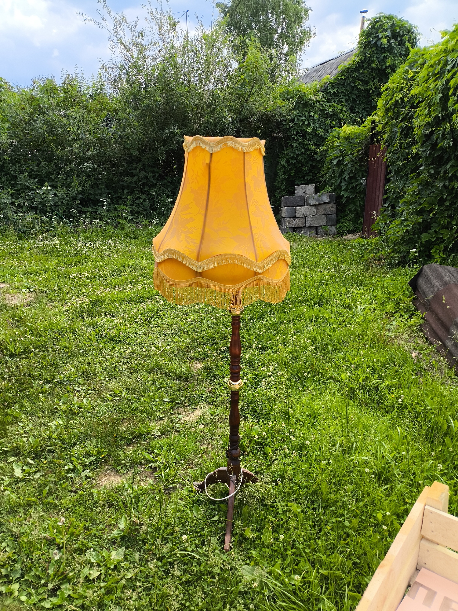 A vintage, ornate floor lamp with a bright orange fabric shade sits in a grassy yard. The lamp has a dark wood pole with decorative brass accents and a fringed shade. The yard is overgrown with grass and small white flowers. In the background, there is a dense green bush, a pile of concrete blocks, a corrugated metal sheet, and the side of a building covered in ivy. The sky is visible and appears partly cloudy. The overall scene suggests a rustic or perhaps forgotten outdoor space.