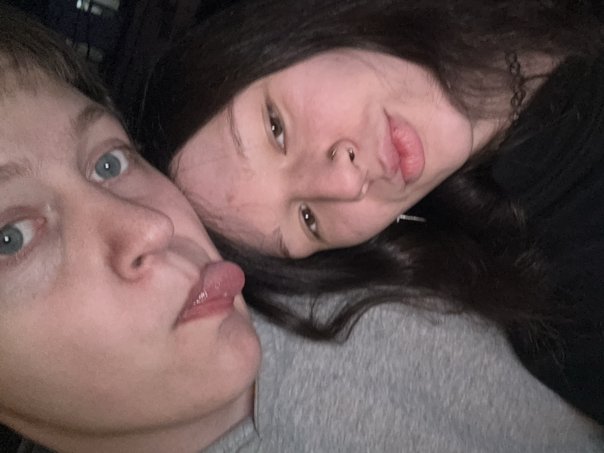 Two people are visible in a close-up, indoor setting. The person on the left has short blonde hair, blue eyes, and is sticking their tongue out. The person on the right has long, dark hair, and a nose piercing. They are close together, suggesting an intimate relationship. One is wearing a grey shirt and the other a black shirt, and there is a patterned necklace around the neck. The location is not discernible and I can't identify it as Saint Petersburg, Russia.