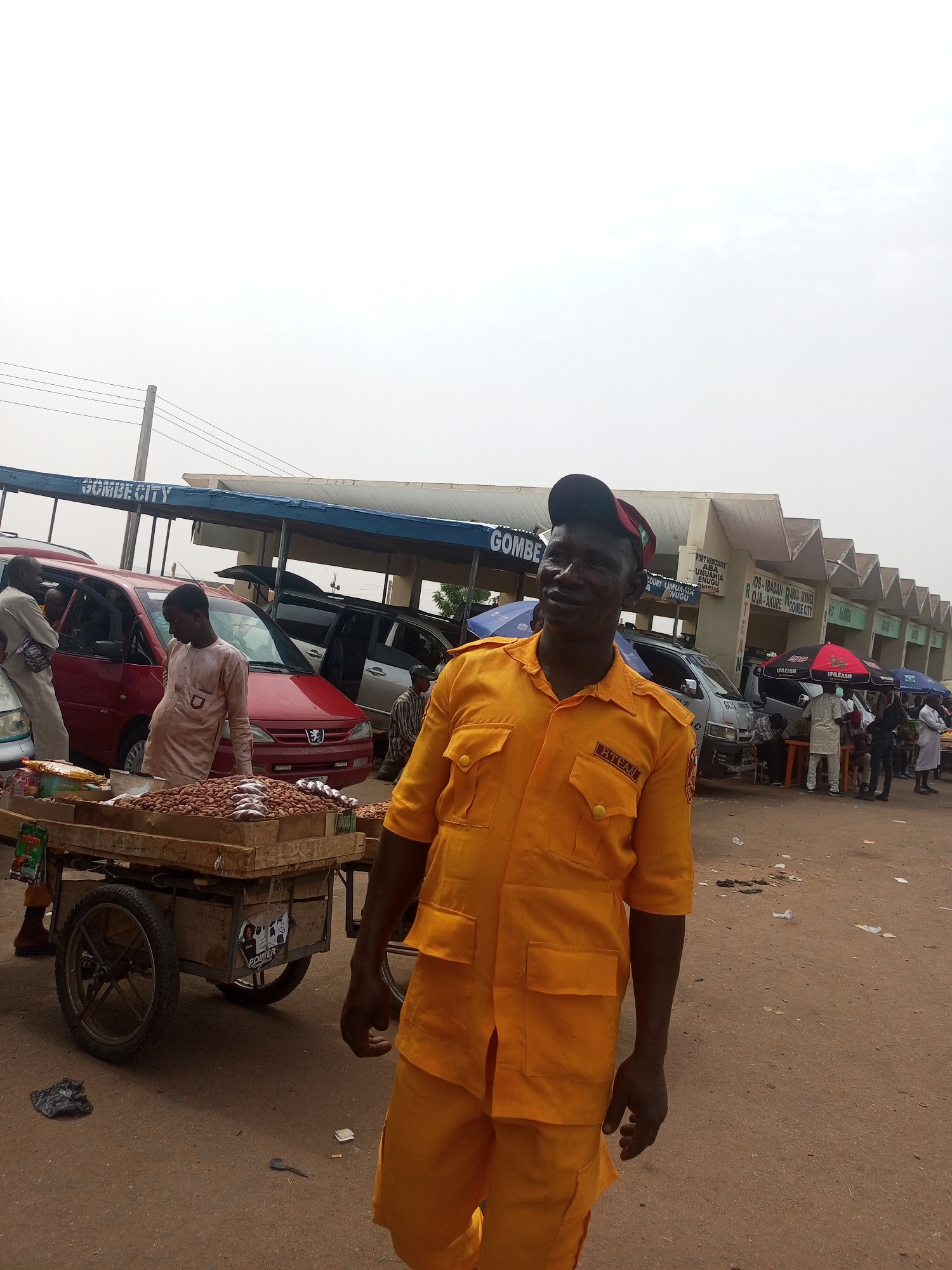 This outdoor photograph captures a bustling scene at a motor park or market in Gombe, Nigeria, under an overcast sky during the daytime. The foreground is dominated by a man in a bright yellow short-sleeved uniform, featuring the text "RTEAN" on his chest pocket, and wearing a dark cap with a red trim. He looks towards the viewer with a slight smile, suggesting a friendly or observant demeanor.
To the left of the main subject, a wooden pushcart, laden with large quantities of groundnuts or similar nuts and some small dried fish, sits on the dusty ground. Another man in a light-colored tunic stands near the cart, close to a red Peugeot sedan parked in the background.
The mid-ground and background reveal more of the transport hub. A long, blue-roofed shelter bears signs that read "GOMBE CITY" and "GOMBE." Further back, a building with a corrugated, wavy roof displays a list of various Nigerian cities, indicating travel destinations such as "PORT HARCOURT," "ABA," "UMUAHIA," "ENUGU," "ONITSHA," "OS-IBADAN," and "AKURE." Several vehicles are parked, and groups of people are visible, some standing under umbrellas marked "UNLEASH." The ground is unpaved and appears dusty, with some discarded items. A sticker on the pushcart also shows the word "POWER." The scene depicts a typical busy day at a local transport and trading center.