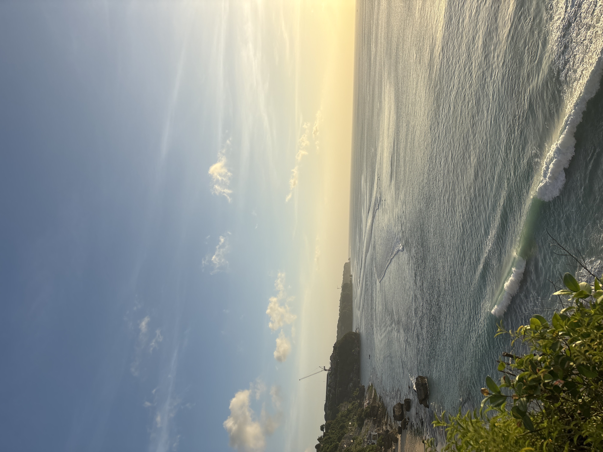 The image captures a coastal view in Kuta Selatan, Indonesia, during the late afternoon or early evening, based on the golden light illuminating the sky and ocean. The horizon line is where the sky and sea meet, with the sun's glow dominating the upper half. A light blue sky speckled with clouds occupies the left side, contrasting with the sun-drenched portion.
The ocean is a deep turquoise color with visible wave patterns. A prominent wave breaks near the bottom right of the frame. The coastline is marked by rocky cliffs covered in dense green vegetation. The cliffs form a jagged edge against the ocean, and buildings and infrastructure, like a crane, are visible along the top of the cliff. The bottom right corner of the image includes green bushes. The photograph provides a wide-angle view of the landscape.