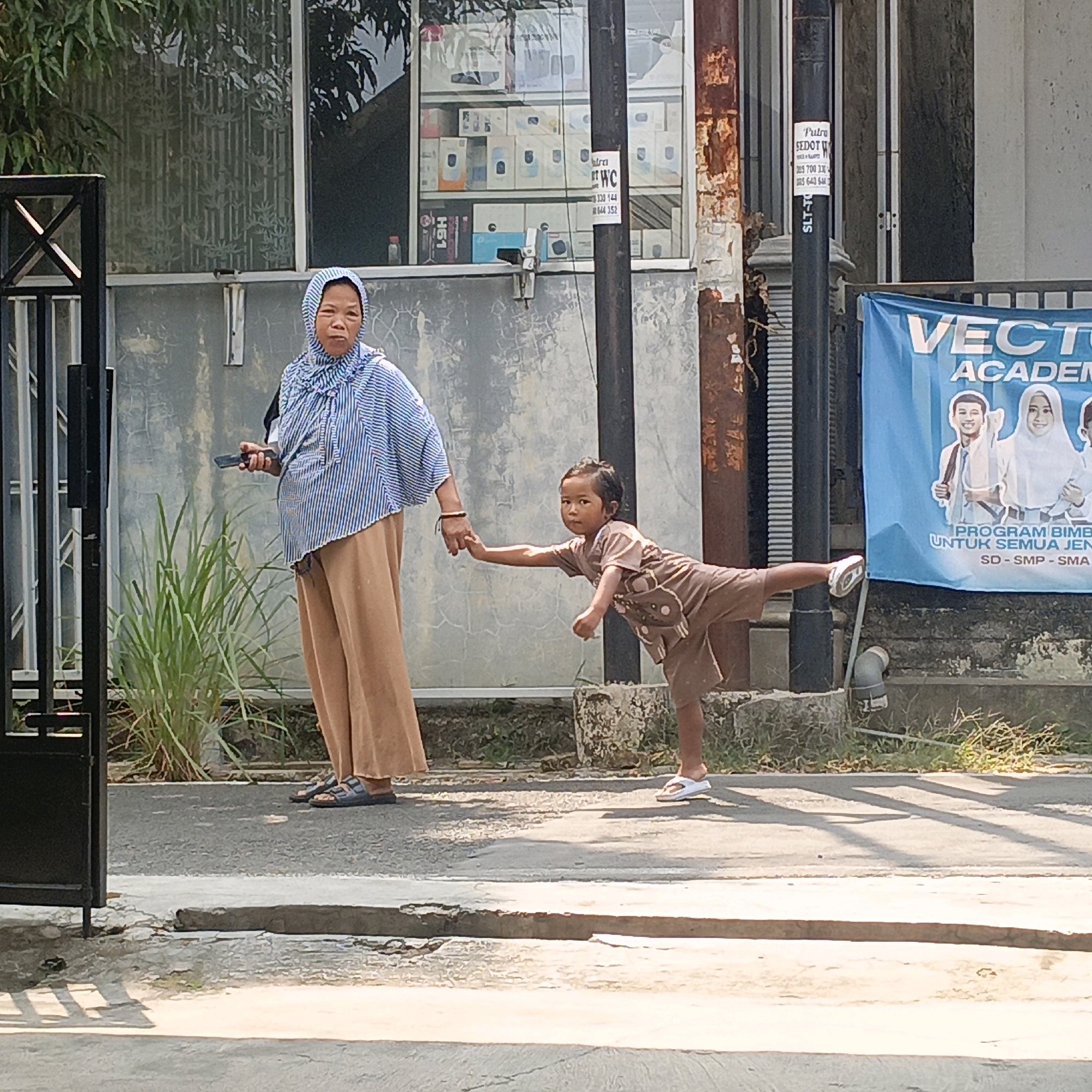 A woman wearing a blue and white striped headscarf and a tan abaya holds the hand of a young child dressed in a tan t-shirt and shorts. The child is balancing on one leg, with the other leg extended backward, and appears to be pulling away from the woman. The woman is holding a black mobile phone in her left hand. They are standing on a paved street in front of a building with a large window displaying various boxes. A blue banner with the text "VECTOR ACADEMY" is visible to the right of the building. The scene is illuminated by sunlight, casting shadows on the ground. The overall setting suggests an outdoor street environment in a commercial area.