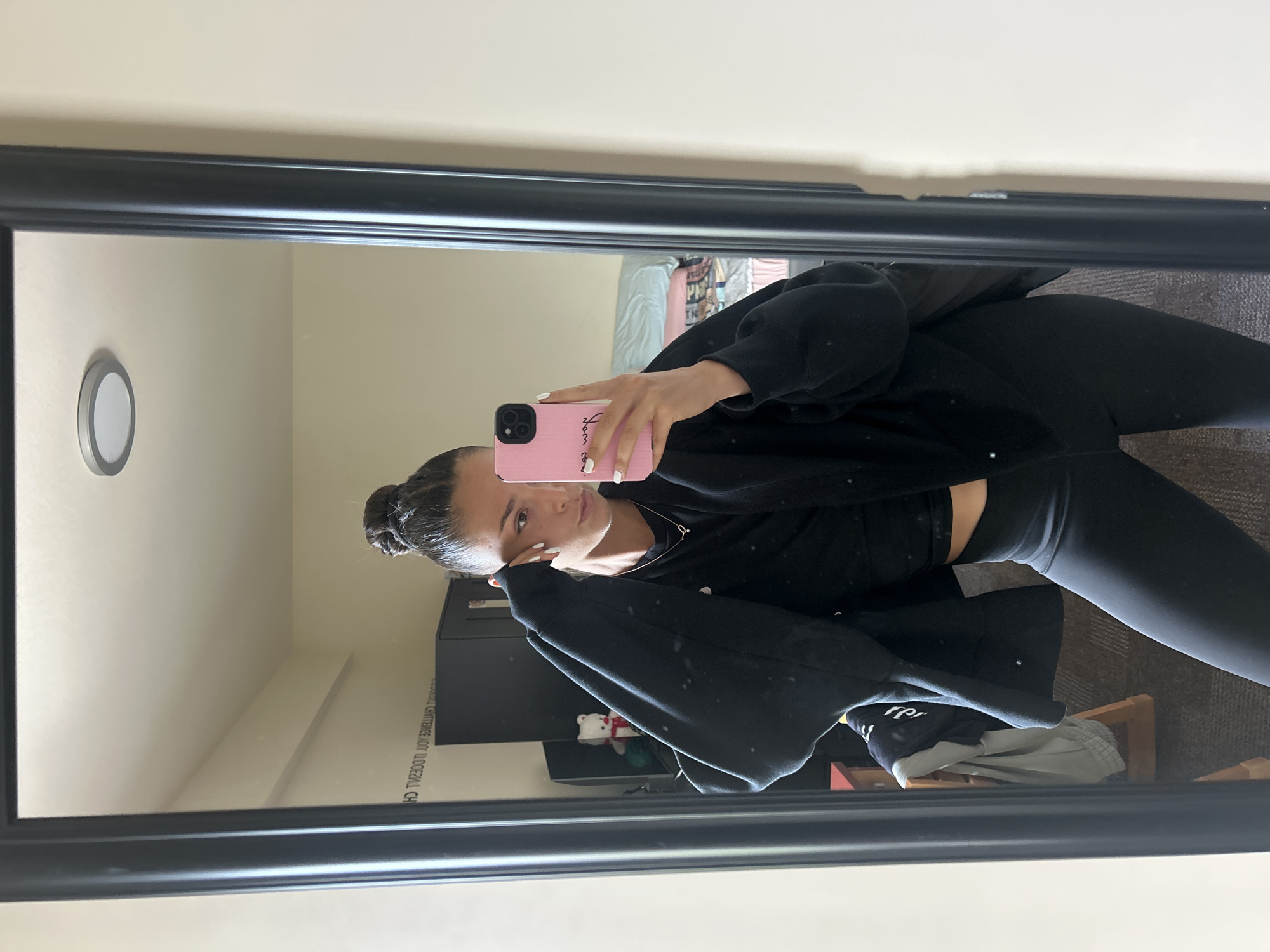 A young woman is taking a selfie in a mirror. She is wearing black leggings, a black sports bra, and a black zip-up hoodie. Her hair is pulled back in a bun. She is holding a pink phone with white nails. In the background, there is a bed, a dresser, a light fixture, and a partially obscured sign written in reverse. The location is the city of University, United States of America.