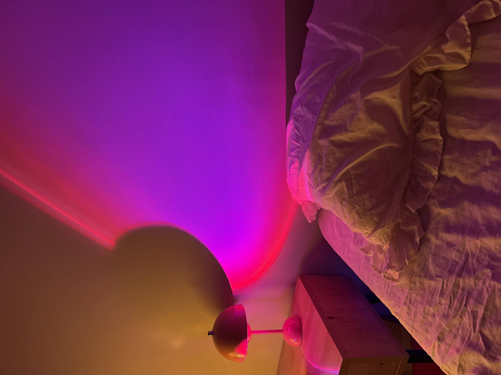 The image shows a bedroom with a bed, nightstand, and a lamp. The room is lit with pink and purple hues, likely from a sunset lamp, casting a circular gradient on the wall and illuminating the bedding and nightstand. The nightstand is made of light-colored wood and holds a pink mushroom-shaped lamp. The bed is unmade with white, ruffled bedding. The scene appears to be an interior shot. There is no indication of Ternopil, Ukraine, or any specific location.