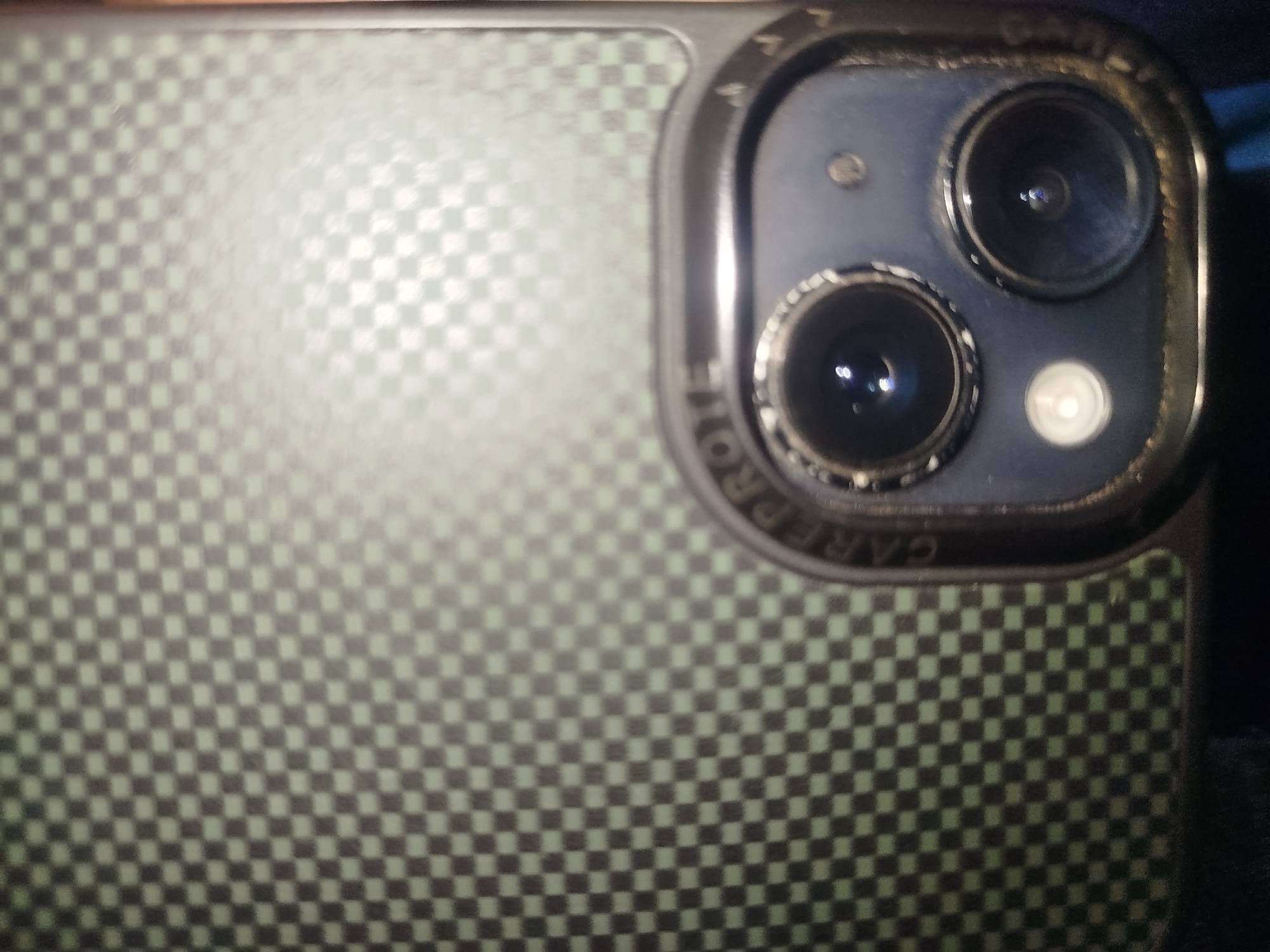 An extreme close-up, slightly blurry, top-down shot focuses on the rear camera module of a black smartphone case with a carbon fiber pattern. The camera module is in the upper right quadrant of the image. The case is a dark gray with a checkered pattern of light green squares. The camera module has a dark blue metal frame with text that is mostly illegible due to blur and reflection, but "CARE" is partially visible. Inside the frame are two circular lenses, a flash, and a small circular sensor to the left of the top lens. The lenses are dark and reflect light. The background is dark and out of focus, suggesting a dimly lit indoor environment. No people or specific city locations are visible.