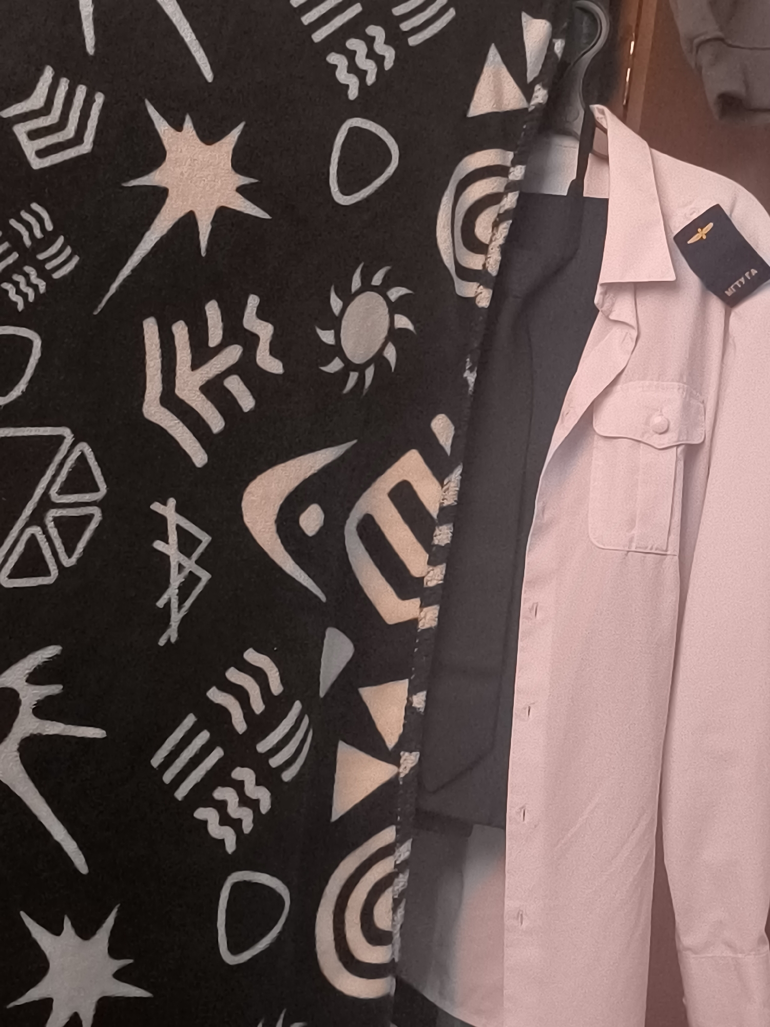 The image shows a close-up view of a light pink button-up shirt hanging on a hanger. The shirt has a pocket on the left breast with a button closure. There is also a dark fabric, possibly a vest or jacket, visible underneath the shirt. On the collar of the shirt, there is a dark blue rectangular patch with gold embroidery, which appears to be an insignia. The patch has a small winged emblem and text that reads "МГТУГА". To the left of the shirt, a black fabric with a pattern of light-colored abstract shapes and symbols is visible, partially obscuring the shirt. This black fabric appears to be a blanket or tapestry. The background is a wall or door, appearing to be a reddish-brown color. There is no clear indication of the location being within the city of Irkutsk, Russia, nor are there any people or specific actions depicted.