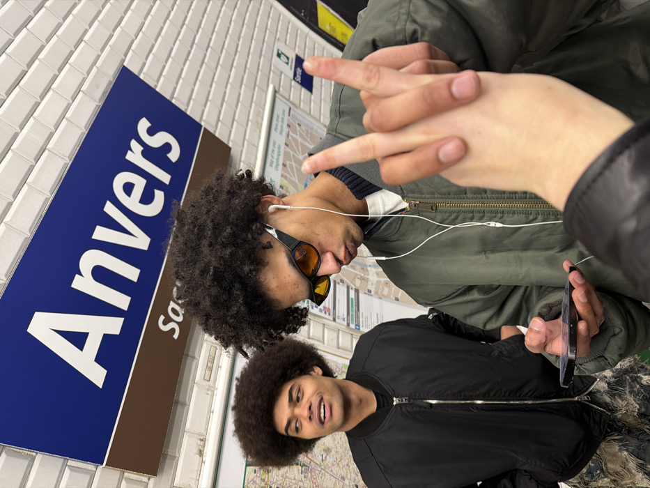 The image depicts an indoor scene at the Anvers Metro station in Paris, France, identifiable by the blue sign displaying "Anvers" and a partial "Sacre Cœur" on a white tiled wall. Two young males with dark, curly afro-style hair are featured in the mid-ground.
The male on the left wears a black bomber jacket and patterned pants. His mouth is open, and he looks towards his right. The male on the right wears an olive green bomber jacket over a white undershirt, black cat-eye sunglasses with orange lenses, and white wired earphones. He holds a black smartphone horizontally in his left hand while making a "duck face" with his lips and raising his right hand in a "V-sign" with extended index and middle fingers. He is looking downwards towards the phone.
A light-skinned hand is visible in the bottom left foreground, partially out of focus, possibly belonging to the individual taking the photograph. The background consists of white rectangular ceramic tiles and blurred subway maps or posters on the wall.