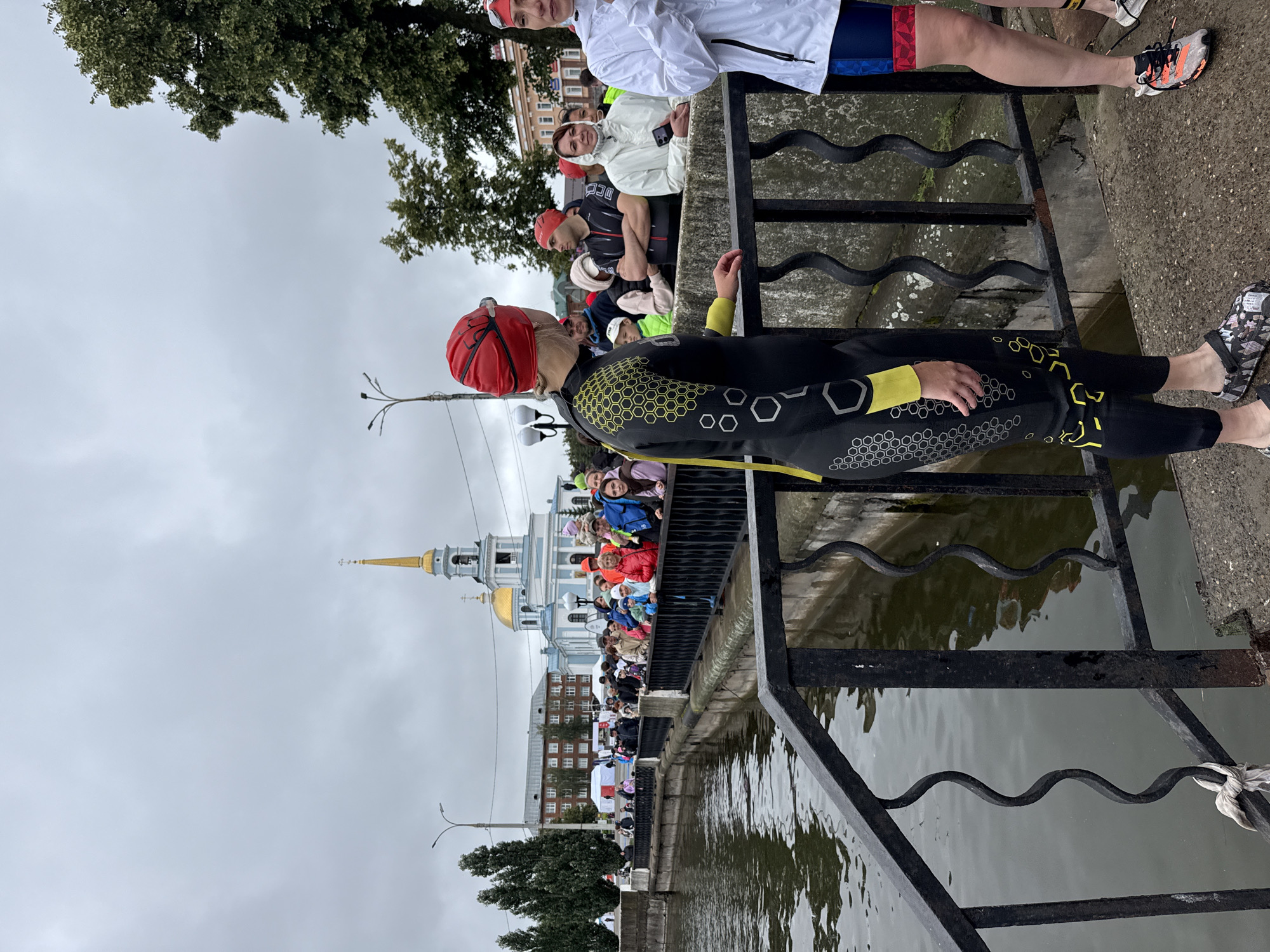 In Votkinsk, Russia, on an overcast day, a group of people stand on a stone embankment beside a canal or river. In the foreground, a woman in a black and yellow wetsuit and red swim cap faces away from the camera, with her left hand resting on a black metal railing. To her right, another woman, seen from the waist down, wears a white jacket and colorful athletic shorts.
Behind them, a crowd of spectators lines the embankment, many holding umbrellas and wearing hats or head coverings. They are looking towards the water. Several white tents are visible in the background, along with trees and buildings. Dominating the skyline is a prominent blue and white church with a golden dome and spire, characteristic of Russian Orthodox architecture. The water is dark and appears calm, reflecting the cloudy sky and surrounding structures. A black metal railing with decorative S-shaped elements runs along the edge of the embankment.