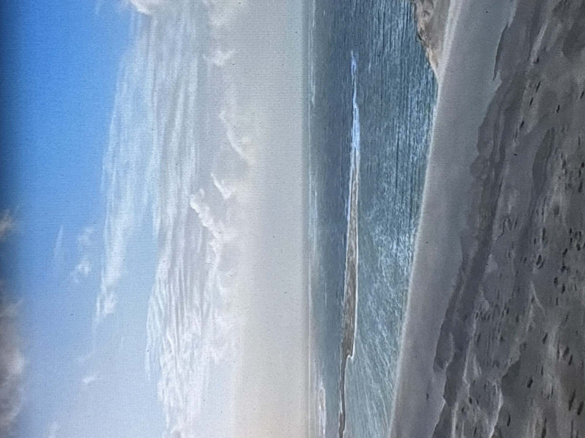 The image depicts a serene coastal landscape with a sandy shore in the foreground and the sea stretching towards the horizon. The water is a clear blue, with gentle waves creating white foam on the surface. A strip of land, possibly an island or a sandbar, breaks the monotony of the sea in the middle distance. The sky is a vibrant blue, dotted with scattered white clouds that create a soft, diffused light. The overall impression is one of tranquility and natural beauty. The location of Zvenigorod, Russia, is not identifiable from the image itself.