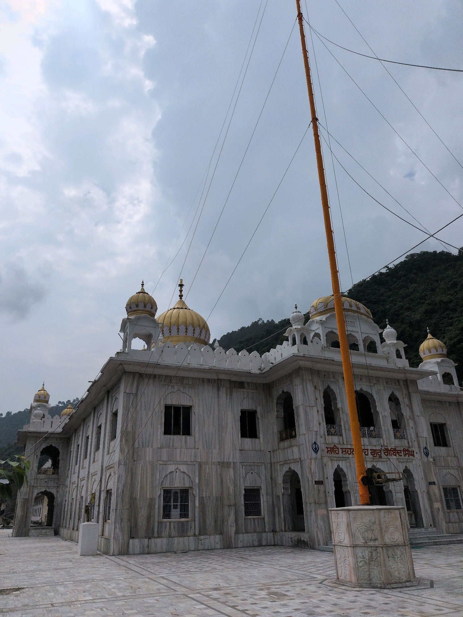The image depicts a Gurdwara in the city of Mandi, India. The Gurdwara is a white marble building with golden domes and intricate carvings. It is surrounded by a paved courtyard and a lush green hillside. A tall, orange flagpole stands in front of the building, adorned with Sikh religious symbols. The sky is cloudy and overcast, suggesting a moderate climate. No people are visible in the image.