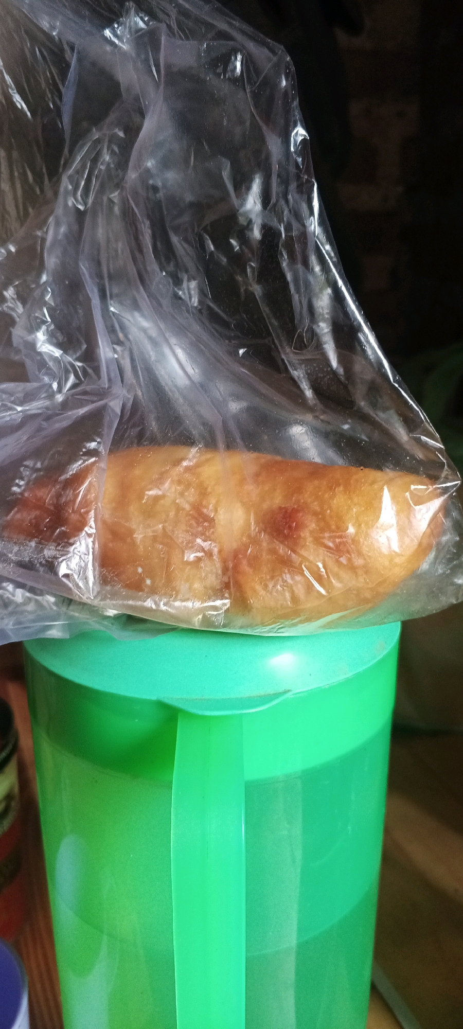 The image shows a baked good, possibly a pastry or bread roll, wrapped in clear plastic. The item is golden brown and appears to be resting on the rim of a bright green plastic container. The background is dark and out of focus, with hints of wooden surfaces. The lighting is artificial and highlights the sheen of the plastic wrap. The scene is indoors, possibly in a kitchen or storage area. There are no people or animals visible in the image. The location in the city of Penanggungan, Indonesia is not specified or inferable from the image itself.
