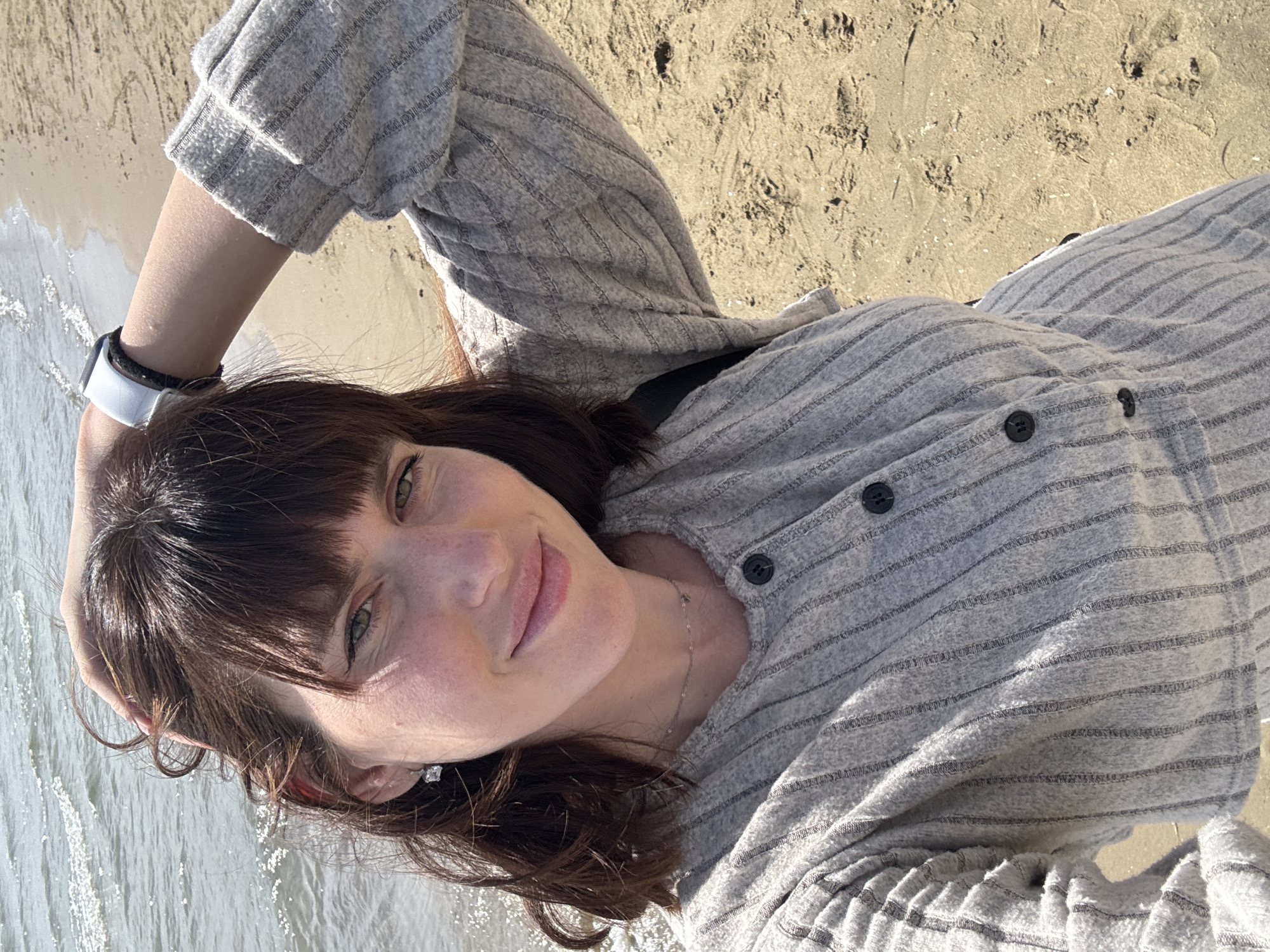 A woman with brown hair and green eyes is lying on a sandy beach next to the water. She is wearing a gray striped, button-down shirt and a white smartwatch on her left wrist. Her head is resting on her left hand. The sand is light beige and appears to be damp near the water. The water appears calm and reflects sunlight. The scene is in Nettuno, Italy.