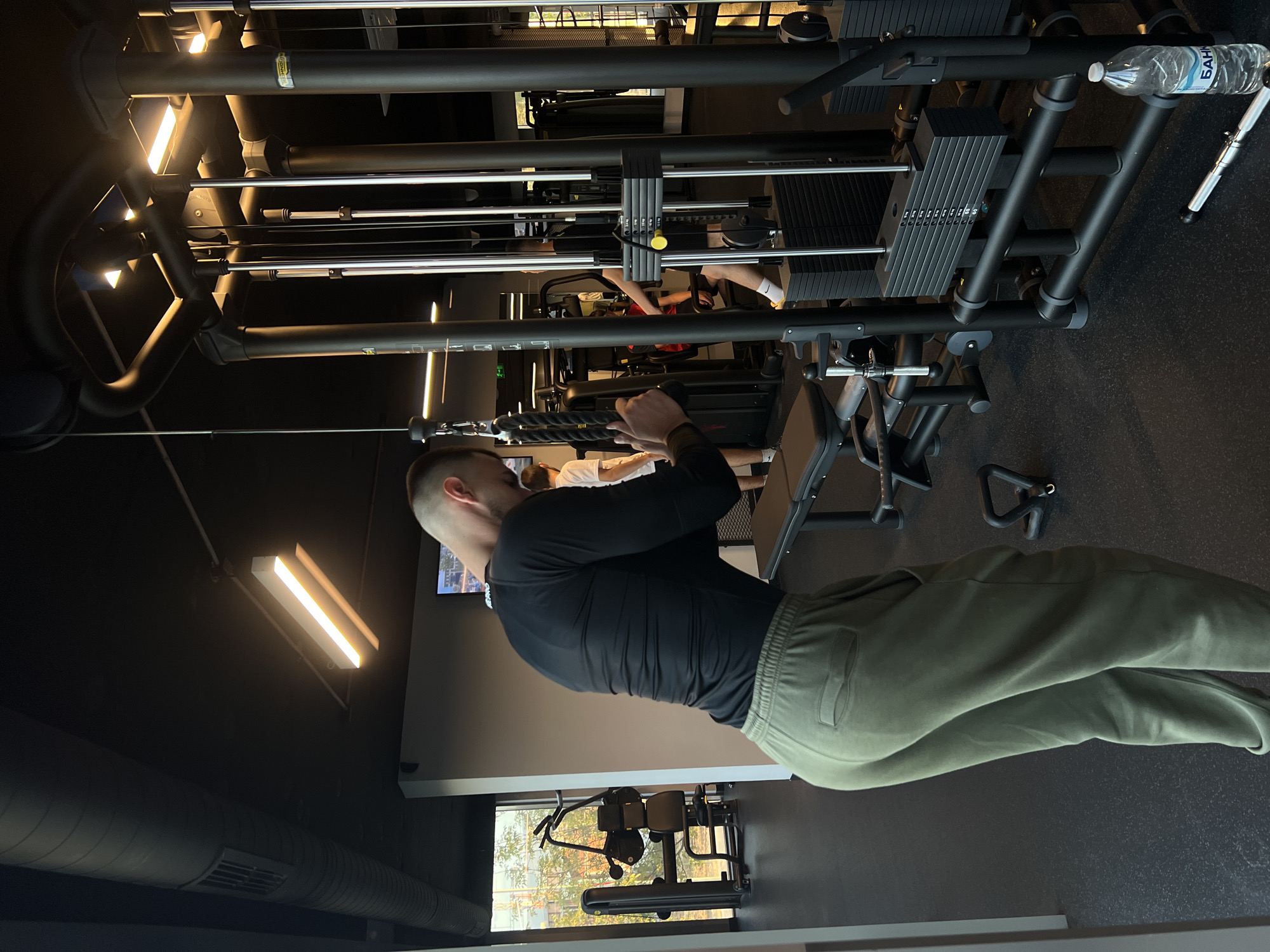 This media file depicts an indoor gym in Plovdiv, Bulgaria, oriented sideways. The main subject is a man, viewed from his back and right side, standing upright and performing an exercise on a cable machine. He wears a long-sleeved black athletic shirt and olive green sweatpants, with his arms bent as he pulls a rope attachment downwards, indicative of a triceps pushdown or similar movement.
The gym features modern fitness equipment with a dark aesthetic. A prominent cable crossover machine occupies much of the frame, showing its intricate structure of metal bars and cables. Several weight stacks are visible on the machine, including one with increment labels (e.g., 10, 20, 30, 40, 50, 60, 70, 80, 90). A transparent plastic water bottle with "БАНКЯ" in Cyrillic text is resting on a part of the machine.
In the background, another individual's legs and lower body are visible, seemingly using another piece of gym equipment. The gym's interior has dark walls and a dark ceiling, from which bright, rectangular fluorescent lights illuminate the space. Large, exposed air ducts also run along the ceiling. A window on the lower left side of the frame offers a glimpse of outdoor foliage and natural light, complementing the artificial lighting. The floor is covered with dark, textured gym flooring. The overall scene conveys a focused workout environment.