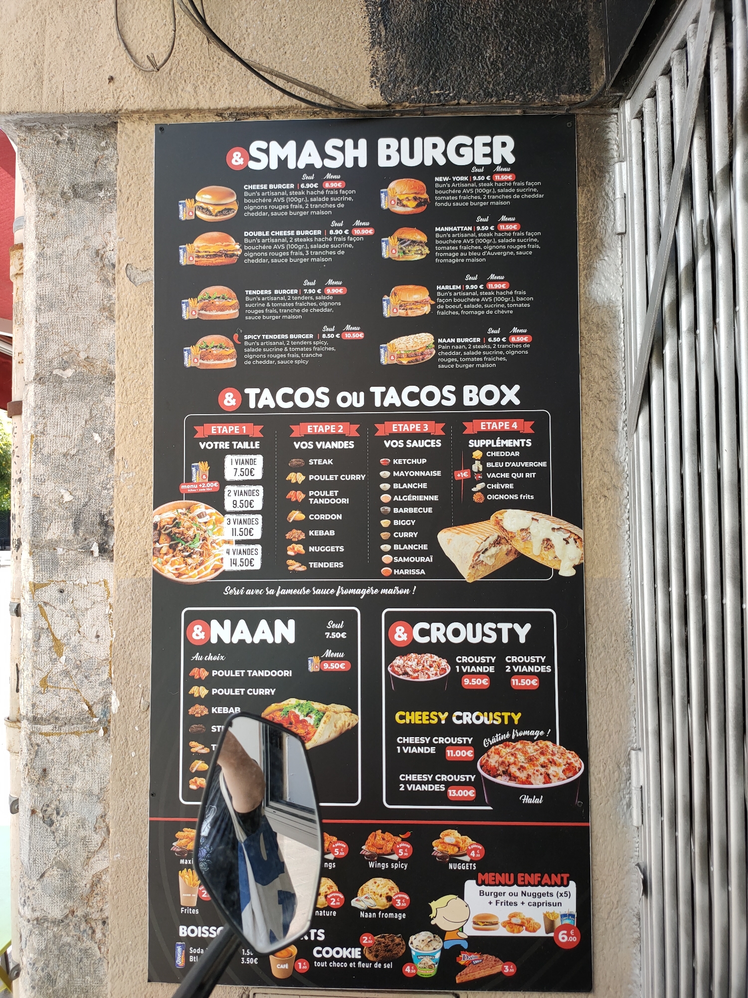 This media file is a photograph taken at an outdoor food stall in Perpignan, France. The main subject is a large, black menu board displaying various burger, taco, and naan options. The menu is divided into sections like "SMASH BURGER," "TACOS OU TACOS BOX," "& NAAN," and "& CROUSTY." Each item is accompanied by a description, price, and often a picture of the food.
The setting appears to be a street-side vendor, with a textured, weathered stone wall on the left and a metal grate structure on the right, suggesting a rustic or urban environment. A motorcycle's side mirror is prominently reflected in the center of the image, showing a person wearing a blue and white t-shirt. This reflection partially obscures the lower section of the menu board, specifically the "NAAN" and "CROUSTY" sections, and some of the food pictures at the bottom.
Visible text includes the names of burger and taco combinations like "CHEESE BURGER," "NEW-YORK," "SPICY TENDERS BURGER," and the process for building a "TACOS BOX" (ETAPE 1-4: VOTRE TAILLE, VOS VIANDES, VOS SAUCES, SUPPLÉMENTS). Prices are clearly listed in Euros (€). Other text indicates "MENU ENFANT" with a price of 6.00€. The overall impression is a vibrant and casual food service operation, likely during daytime given the lighting.