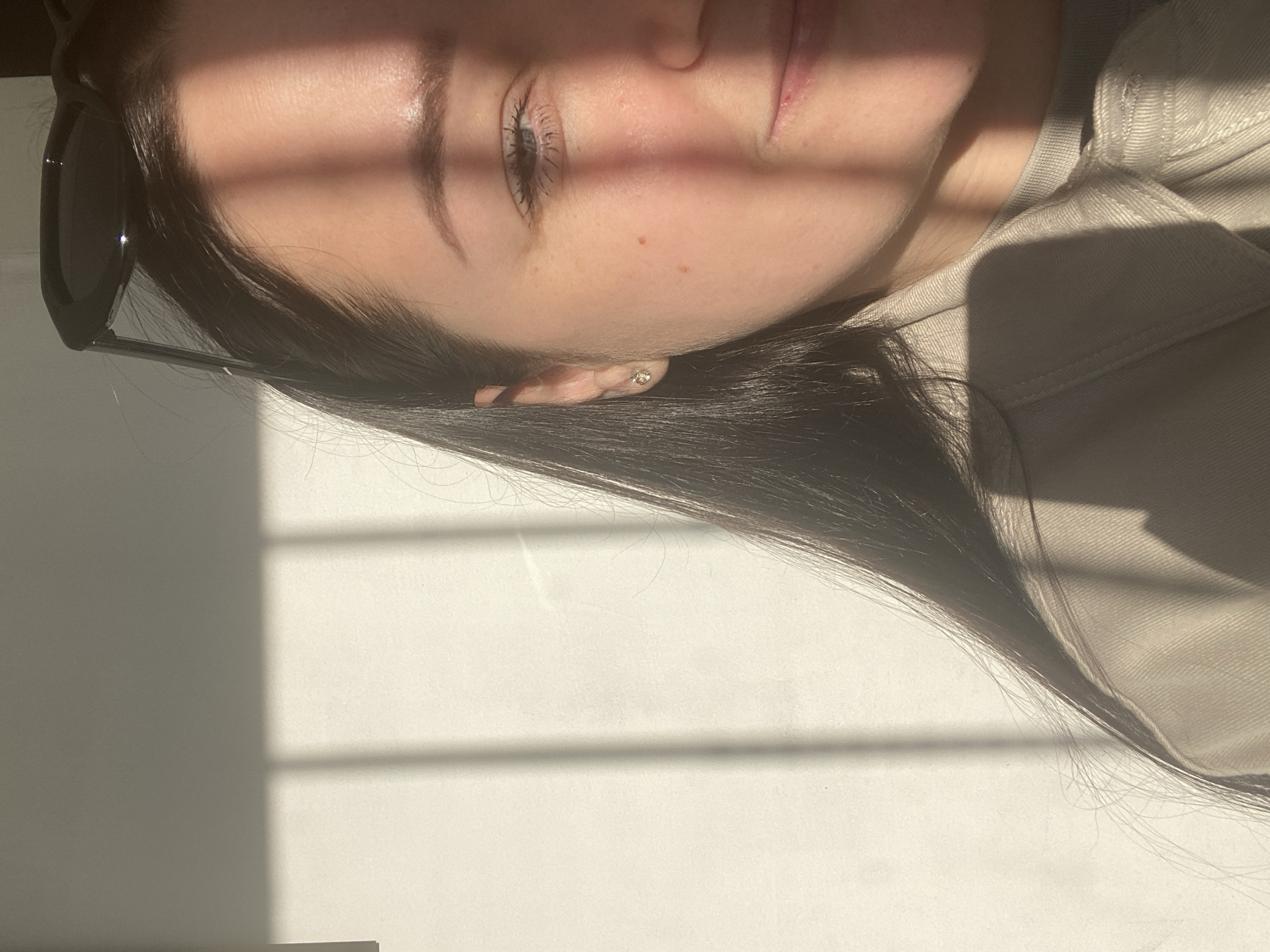 The close-up image presents a rotated portrait of a young woman, captured in Sochi, Russia. Her head is tilted, with long, dark hair flowing towards the bottom of the frame. She wears black sunglasses resting on her forehead, and a small, sparkly stud earring is visible in her right ear. Her expression appears calm, with her right eye open and looking subtly upwards. She is dressed in a light-colored, possibly beige or light grey, textured jacket or shirt.
Strong, direct sunlight streams into the indoor setting, creating prominent, geometric shadows across her face, hair, and the plain light-colored background. These shadows suggest the presence of window bars or blinds, and indicate a sunny day, likely during the morning or late afternoon when the sun is lower in the sky. The interplay of bright highlights and deep shadows creates a high-contrast and somewhat pensive mood. No text is visible in the media.