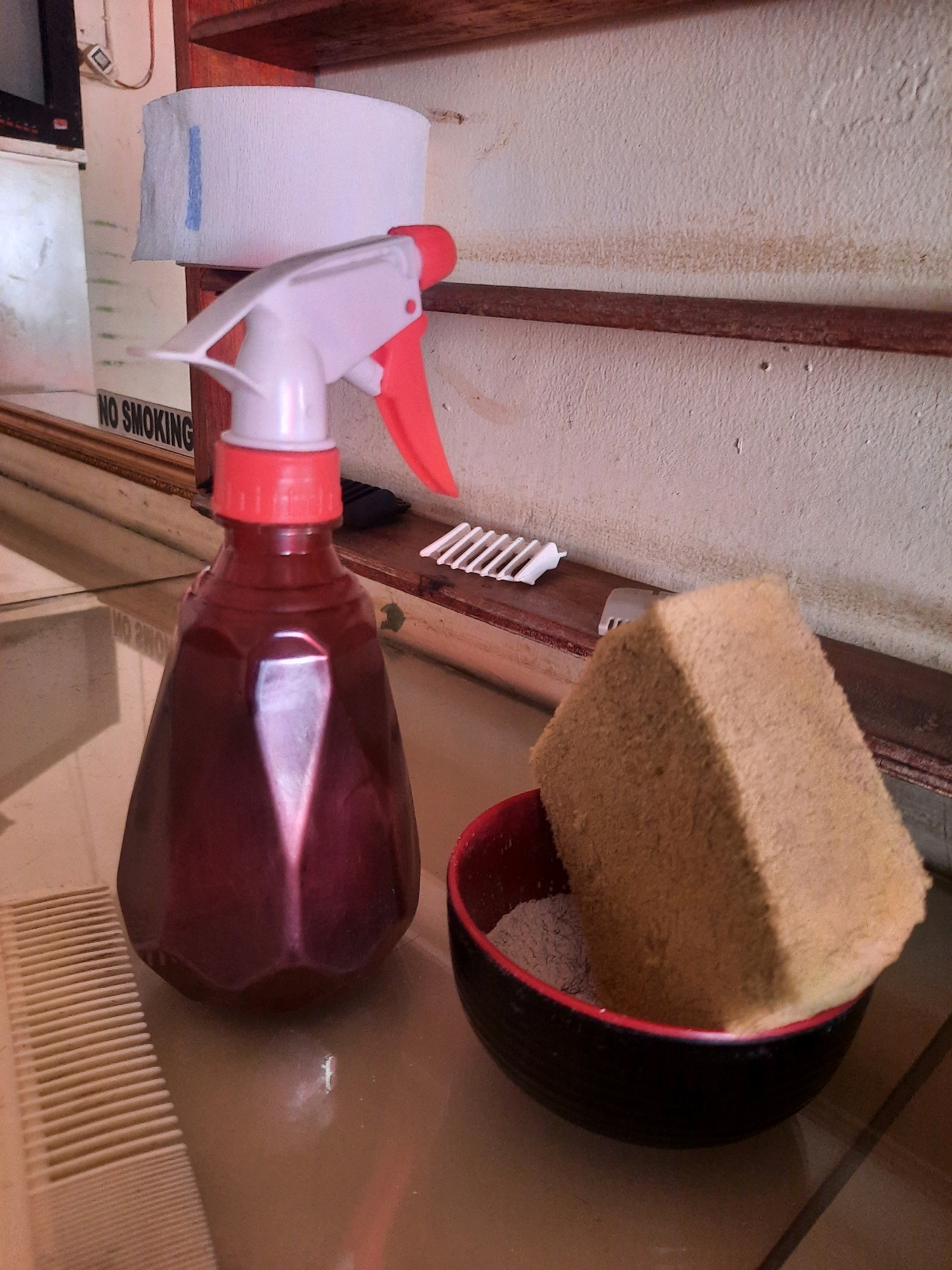 This is a close-up, eye-level shot of cleaning supplies on a surface. On the left is a dark red spray bottle with a white and red spray nozzle. In front of the bottle is a light-colored plastic comb. To the right of the spray bottle, a light brown, rectangular sponge sits in a black bowl with a red rim. The bowl appears to contain a gray powder. Behind the spray bottle, a roll of white paper towels is visible, attached to a wooden shelf. A "NO SMOKING" sign is partially visible behind the spray bottle. The background is a textured, off-white wall. The scene is likely indoors, possibly in a kitchen or bathroom in East Jakarta, Indonesia, given the context of cleaning supplies.