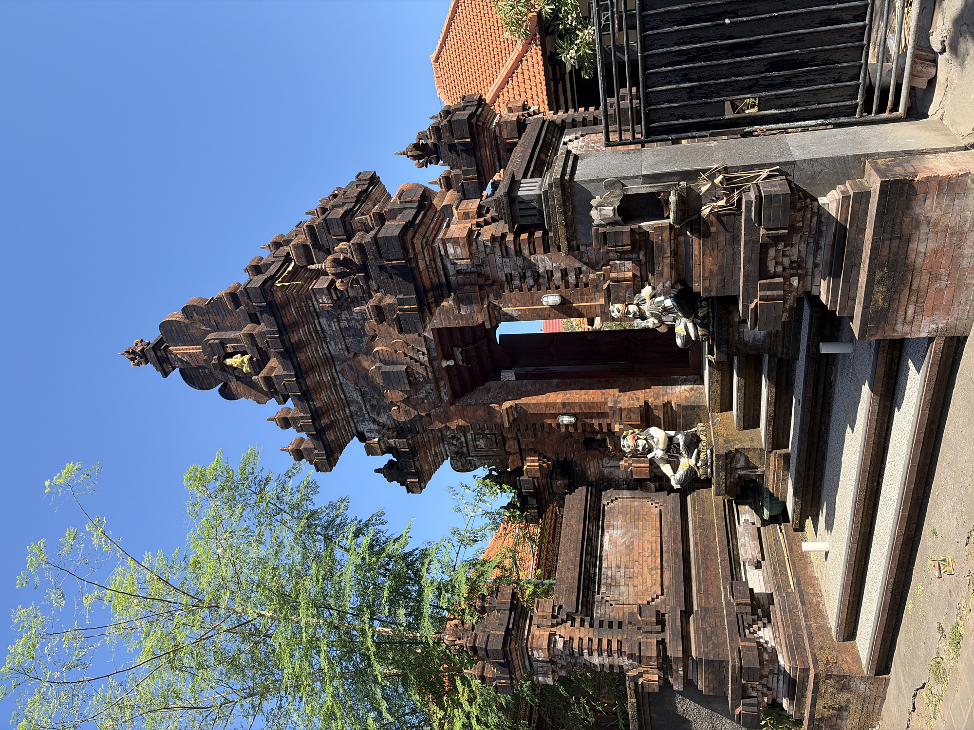 A balinese temple entrance gate in Kuta Selatan, Indonesia. The gate is constructed of dark red brick with intricate carvings and several tiered roof structures. Two seated guardian statues, one on each side of the stairs, flank the entrance. One statue is wearing a red and white headpiece and a black and white striped garment. The other statue is wearing a floral headpiece and a black and white outfit. A tall green tree with delicate branches is visible to the left of the gate, and a red tiled roof is visible behind the gate. A black metal gate is to the right of the entrance. The sky is clear and blue.