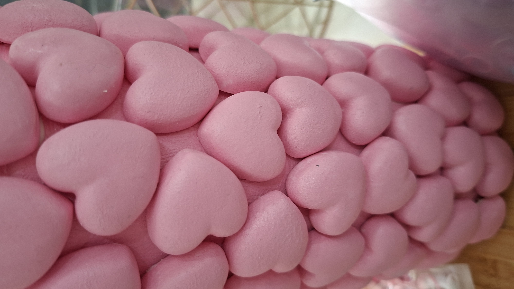 A close-up photograph shows a textured surface covered with numerous small, three-dimensional pink heart shapes. The hearts are densely packed together, creating a repetitive pattern across the field of view. The shapes appear to be made of a somewhat porous or textured material. The light source is from the top right, creating shadows on the left side of each heart. Some parts of the image are slightly out of focus, particularly towards the right edge. There is no visible information to determine the location in the city of Chaikovsky, Russia. There are no people or actions depicted.