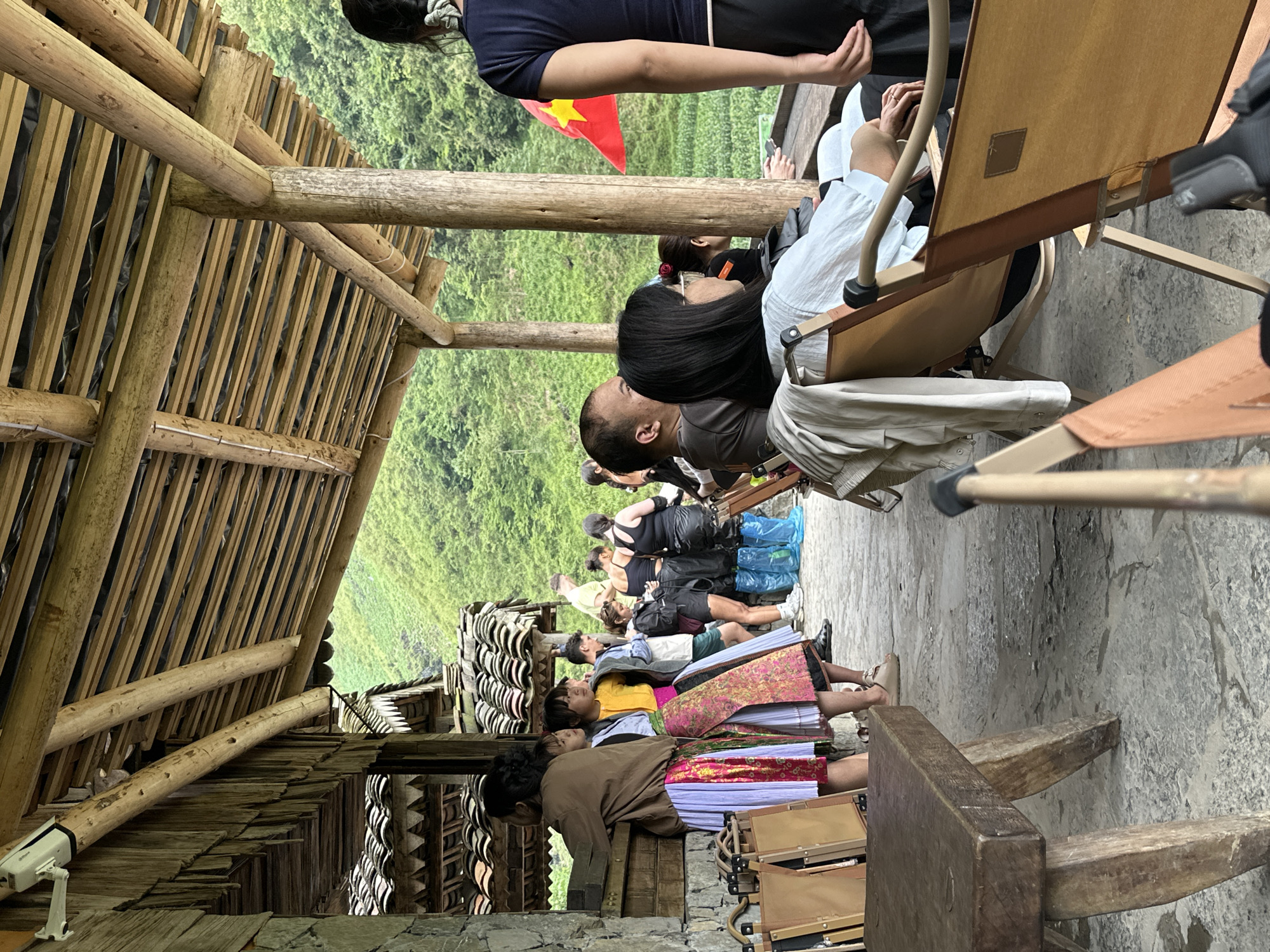 The image shows a group of people, including Vietnamese people in traditional dress, resting on a stone-paved ground under a wooden structure with a bamboo roof, possibly in Yên Minh Commune, Vietnam, given the traditional clothing. The people are seated in chairs or on the ground. A Vietnamese flag is visible in the background of lush green mountains. A security camera is also visible near the structure.