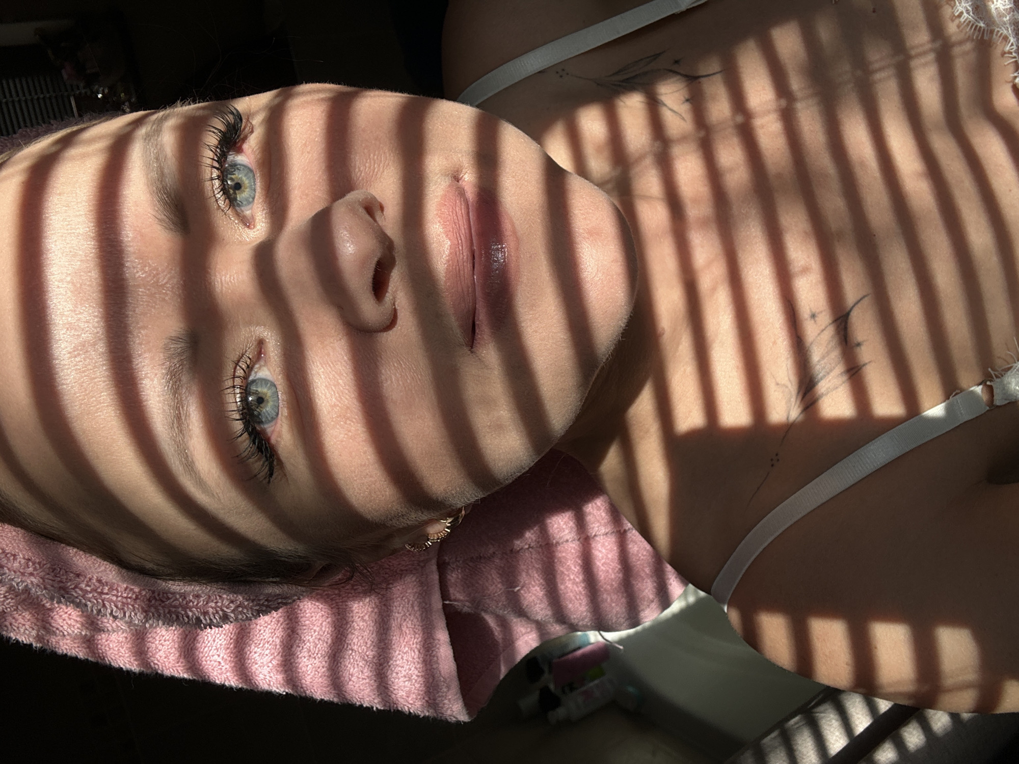 A fair-skinned person lies on a pink towel, seemingly indoors. Light from a window blind casts parallel shadows across their face and upper chest. The person has blue eyes, long eyelashes, and light pink lips. Tattoos are visible on their chest above a white bra strap. The setting is potentially residential. The location (Chaikovsky, Russia) is not apparent from the image itself.