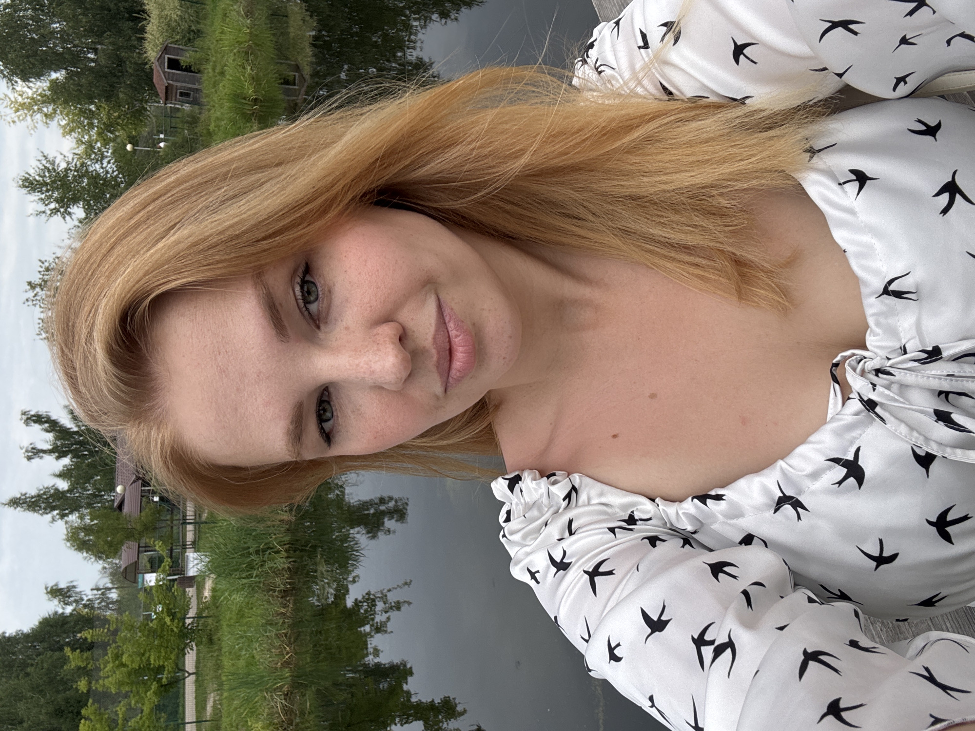 A young woman with blonde hair and freckles is taking a selfie outdoors. She is wearing a white top with black bird patterns. She is standing on a wooden surface, possibly a deck, with a body of water and lush greenery behind her. Trees, reeds, and a small wooden structure are visible in the background, suggesting a park or natural area. The sky is overcast. The scene appears to be a serene, natural setting within the city of Yaroslavl, Russia, as indicated by the mention of its location.