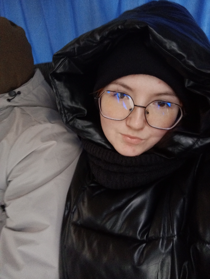 A selfie-style portrait features two individuals indoors against a solid blue, textured background, possibly a fabric curtain or upholstery. The primary subject, a young adult female, occupies the right half of the frame, looking directly at the camera with a neutral expression. She wears a glossy black puffer jacket with an oversized, padded hood pulled up, a black knitted beanie underneath, and a black ribbed neck gaiter. Her hexagonal-framed glasses exhibit prominent blue light reflections on the lenses. Partially visible on the left side of the frame is the upper torso and head of a second individual, dressed in a light grey or beige puffer jacket with a dark brown/olive-colored hood or cap partially visible above it. The precise location within the city of Tap'iushka, Russia, cannot be determined from the visual information provided.