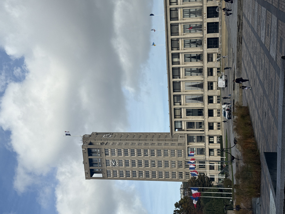 This image captures the Hôtel de Ville (City Hall) complex in Le Havre, France, specifically in the Place de l'Hôtel de Ville, under a partly cloudy sky. The dominant structure is a tall, rectilinear concrete clock tower, approximately 20 stories high, featuring multiple clock faces on its visible sides and numerous evenly spaced, recessed rectangular windows. A French tricolor flag flies from a pole atop the tower.
Adjacent and to the right of the tower is the lower, wider, multi-story main Hôtel de Ville building, constructed in a modernist style with a concrete facade, large recessed window bays, and vertical architectural elements. The inscription "HOTEL DE VILLE" is visible above the ground floor entrance area. On the roof of this building, several flags are displayed on flagpoles, including a blue and yellow Ukrainian flag and other unidentified flags. To the left of the main building, at a lower elevation on the plaza, a series of flagpoles display multiple French tricolor flags.
The foreground and midground consist of a large, paved urban plaza with a geometric pattern of light and dark rectangular tiles. In the immediate foreground, a rectangular planter or water feature contains dry, brown vegetation and several dark, abstract, bird-like metallic sculptures.
Pedestrian activity is observed across the plaza. In the lower right quadrant, a woman wearing dark clothing walks with a small child, who is on a scooter or similar wheeled toy, both moving away from the viewer. Further back, on the plaza near the main building's entrance, two white utility-style vans are parked. Several individuals are gathered around these vehicles, some appearing to be engaged in work or conversation. Additional scattered pedestrians are visible walking across the plaza.