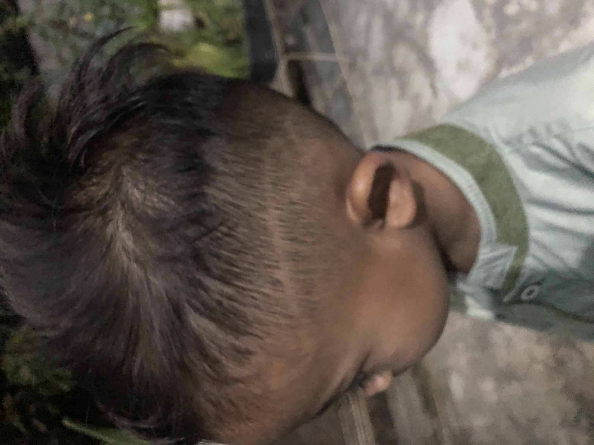 The media file is a close-up, sideways shot of a young child's head, focusing on their hair and ear. The child appears to have dark, short hair styled with a fade or undercut, revealing lighter skin beneath. They are wearing a light blue or green shirt with a green and white striped collar. The background is blurred, showing hints of green foliage and a grey, textured surface, possibly a wall or pavement. The lighting suggests it might be daytime, but the overall image is somewhat grainy and dark, making precise details difficult to discern. The child's expression is not visible as their face is turned away. There is no visible text in the image. The setting appears to be outdoors, given the presence of greenery, and the context provided places the location in Tenggilis Rejo, Indonesia.