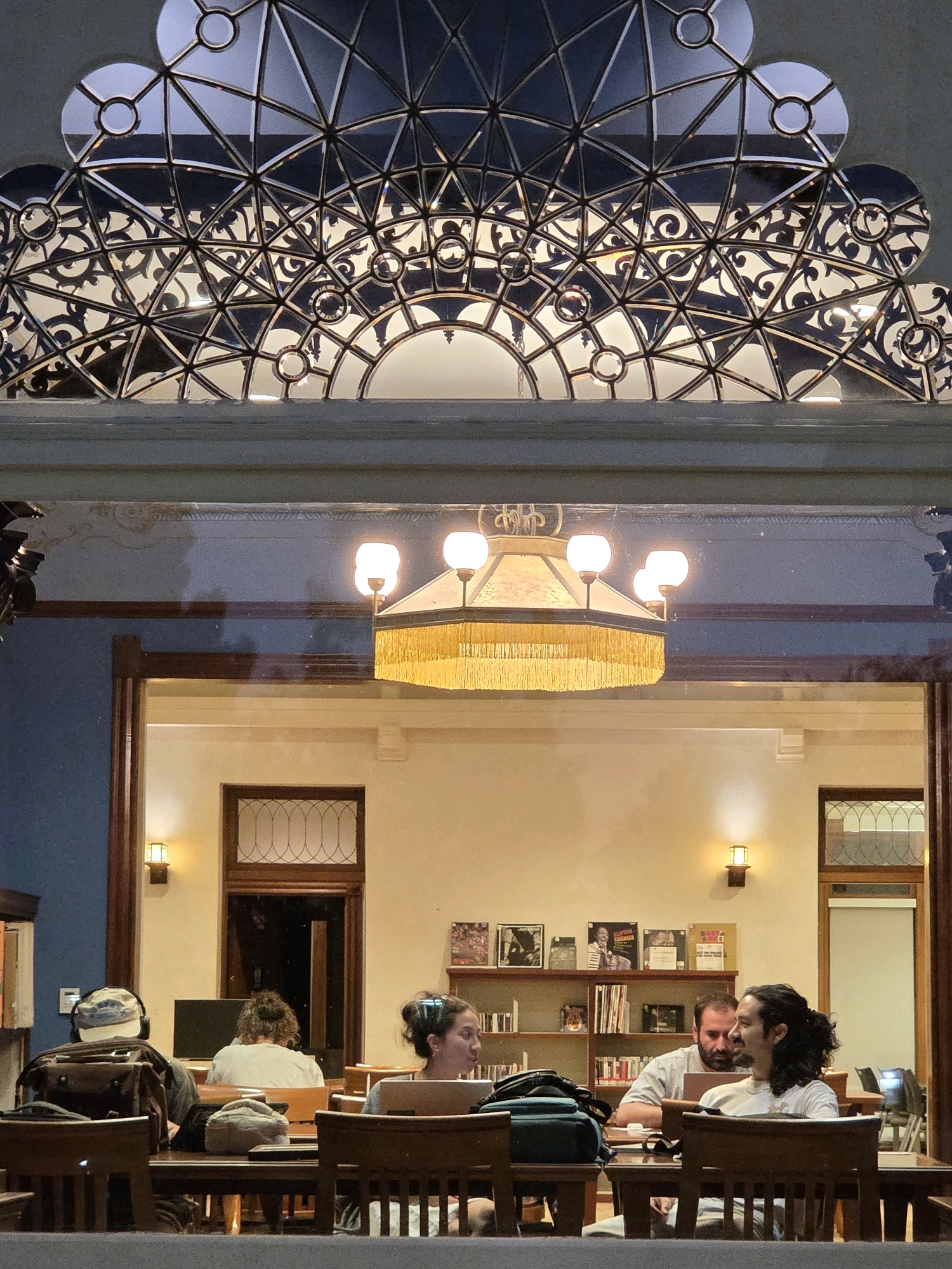 The image captures an interior view of what appears to be a library or study hall in Glendale, United States of America. The scene is illuminated by a prominent, ornate chandelier with a golden fringe and six globes, suspended from the ceiling. Above the chandelier, a decorative geometric pattern of metal and glass is visible.
In the foreground, four individuals are seated at wooden tables, engaged in various activities. On the left, a person wearing headphones and a patterned cap is facing away from the camera, with a backpack resting on their chair. Next to them, another person is seated with their back to the viewer, looking towards a computer screen. To the right, two individuals are facing each other across a table. One person, with dark curly hair, is looking to their right and smiling, while the other person is looking down at their laptop.
Behind the seated individuals, a bookshelf filled with books and framed pictures is visible. Another wooden door with decorative glass panels is present in the background, along with wall-mounted lights. The overall ambiance suggests a quiet and studious environment. The reflection on the glass in the foreground indicates the image was taken from outside the room, looking in.