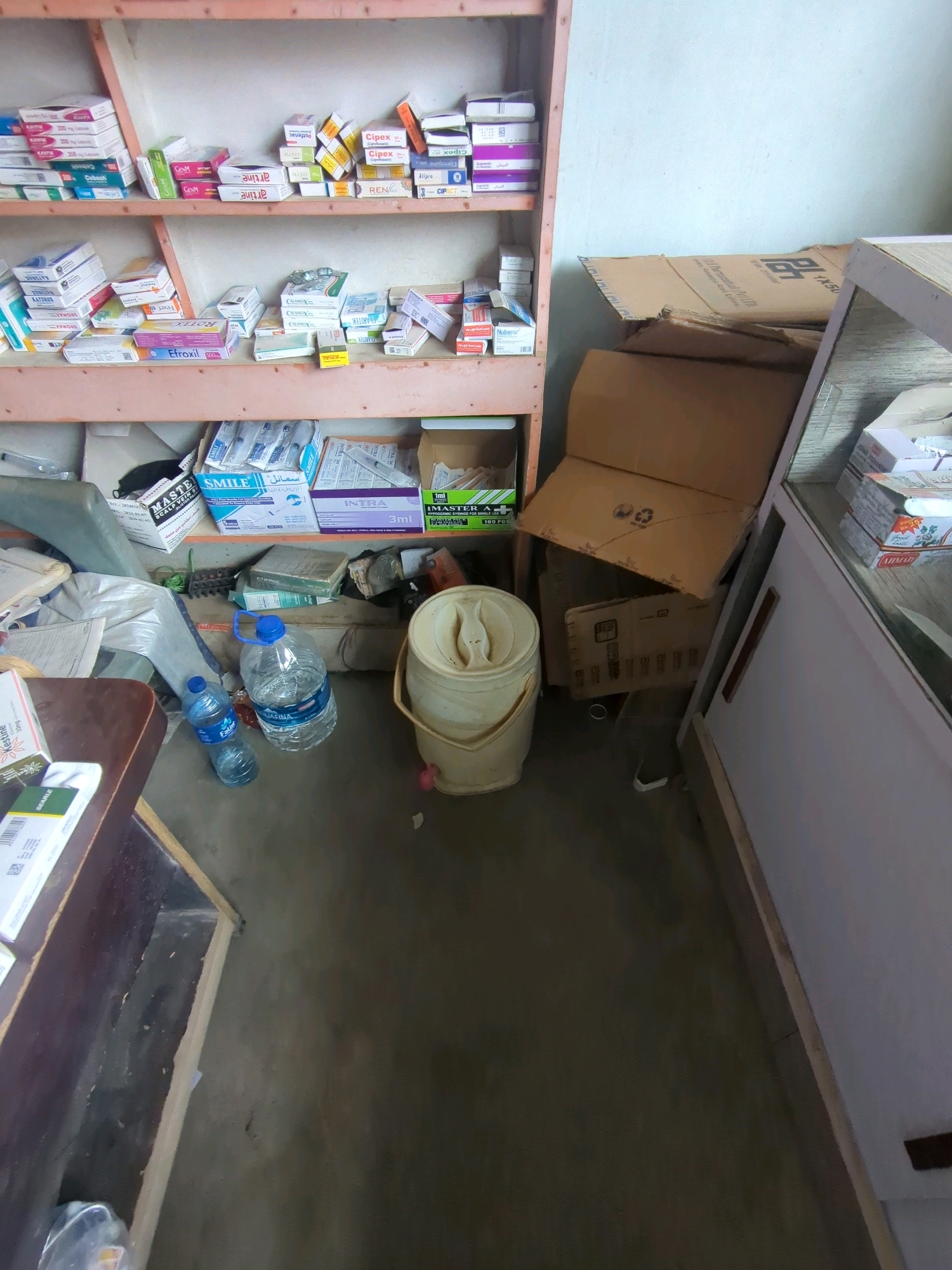 The image depicts the interior of what appears to be a small pharmacy or medical supply store in Karachi Division, Pakistan. The scene is cluttered with various medical products, predominantly pharmaceuticals in boxes and packaging.
On shelves, a wide assortment of medicines is organized, including boxes with labels like "KAYFEX," "Cipla," "Alipro," and "INTRA 3ml." There are also packages of syringes visible on one of the shelves. Beneath the shelves, the floor is covered with more items, including cardboard boxes, plastic bags, and two large bottles of water. One bottle is partially obscured, and the other is a large Aquafina bottle with a blue cap. To the right, there is a beige plastic bucket with a lid and a handle, and a light-colored, possibly refrigerated, display case. Cardboard boxes are stacked in the middle ground, some appearing to be shipping or storage containers. The overall environment suggests a functional but not meticulously organized retail space. The lighting appears to be natural, coming from an unseen source, and illuminates the cluttered environment.