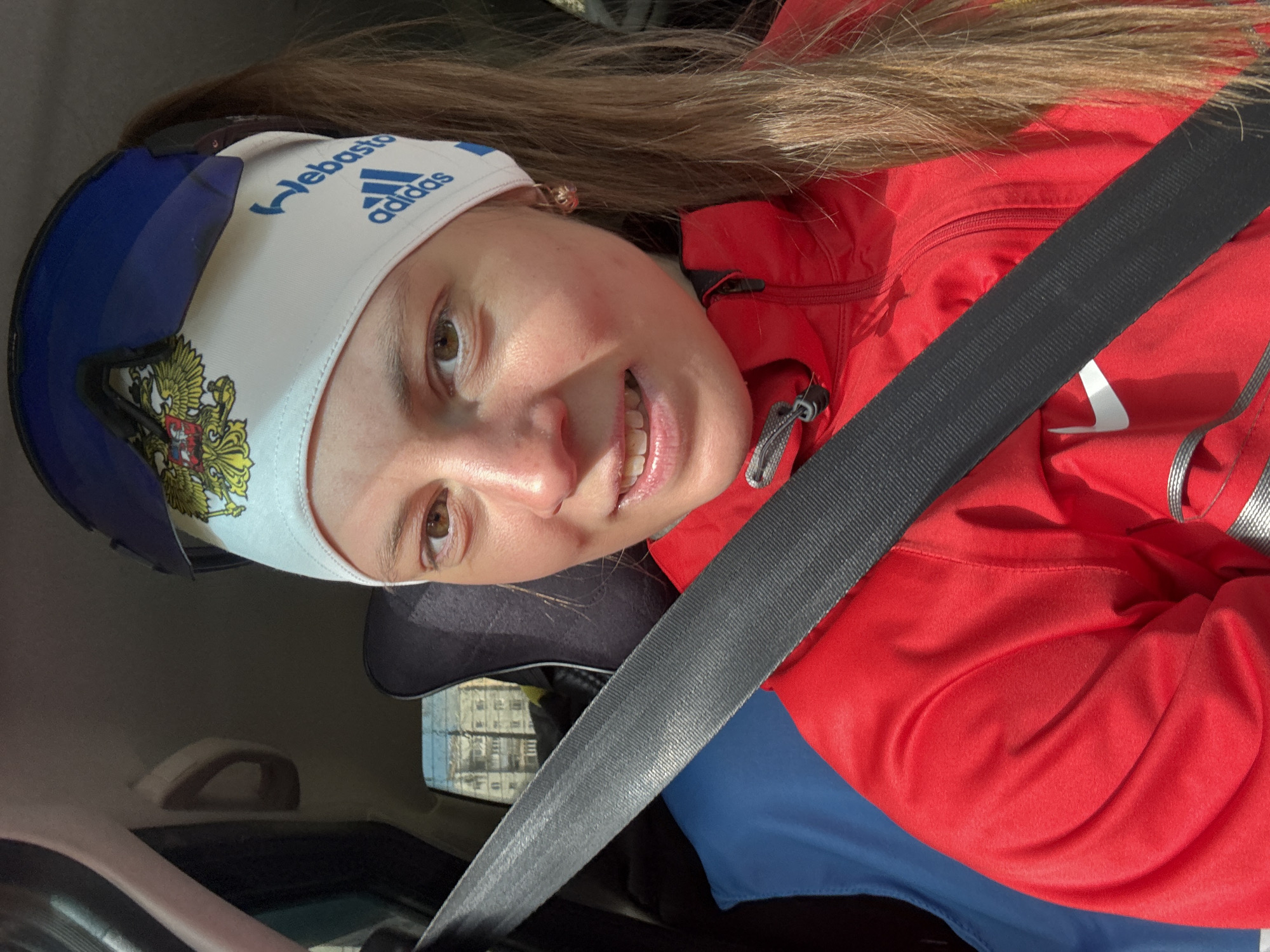A woman is in a car, wearing a red jacket and a white headband with "Webasto" and an Adidas logo on it. She has a seatbelt fastened across her torso. The headband also features the Russian coat of arms. She has brown hair and light skin. Outside the car window, there is a building with multiple floors. The image appears to be taken inside a car in Chaikovsky, Russia based on the emblems and writing on her headband.