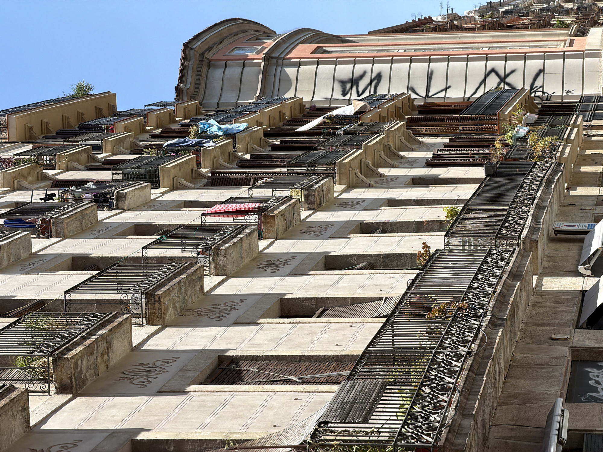 The image displays the facade of an old building in Barcelona, Spain, likely in a historic district. The building features multiple floors with balconies on each level. The balconies are adorned with ornate black metal railings and supported by stone brackets. Some balconies have potted plants, while others have laundry hanging out to dry, including a red and white striped towel, blue and white fabric, and a white sheet. The building's exterior walls are a light beige color with decorative patterns. Numerous windows with closed wooden shutters are visible. The facade of an adjacent building, which is pink and cream colored with a curved corner and a rounded roof, is also partially visible. The sky above is clear and blue. The sunlight casts sharp shadows on the building's facade, indicating a bright day. The ground level of the building has shop fronts with awnings.