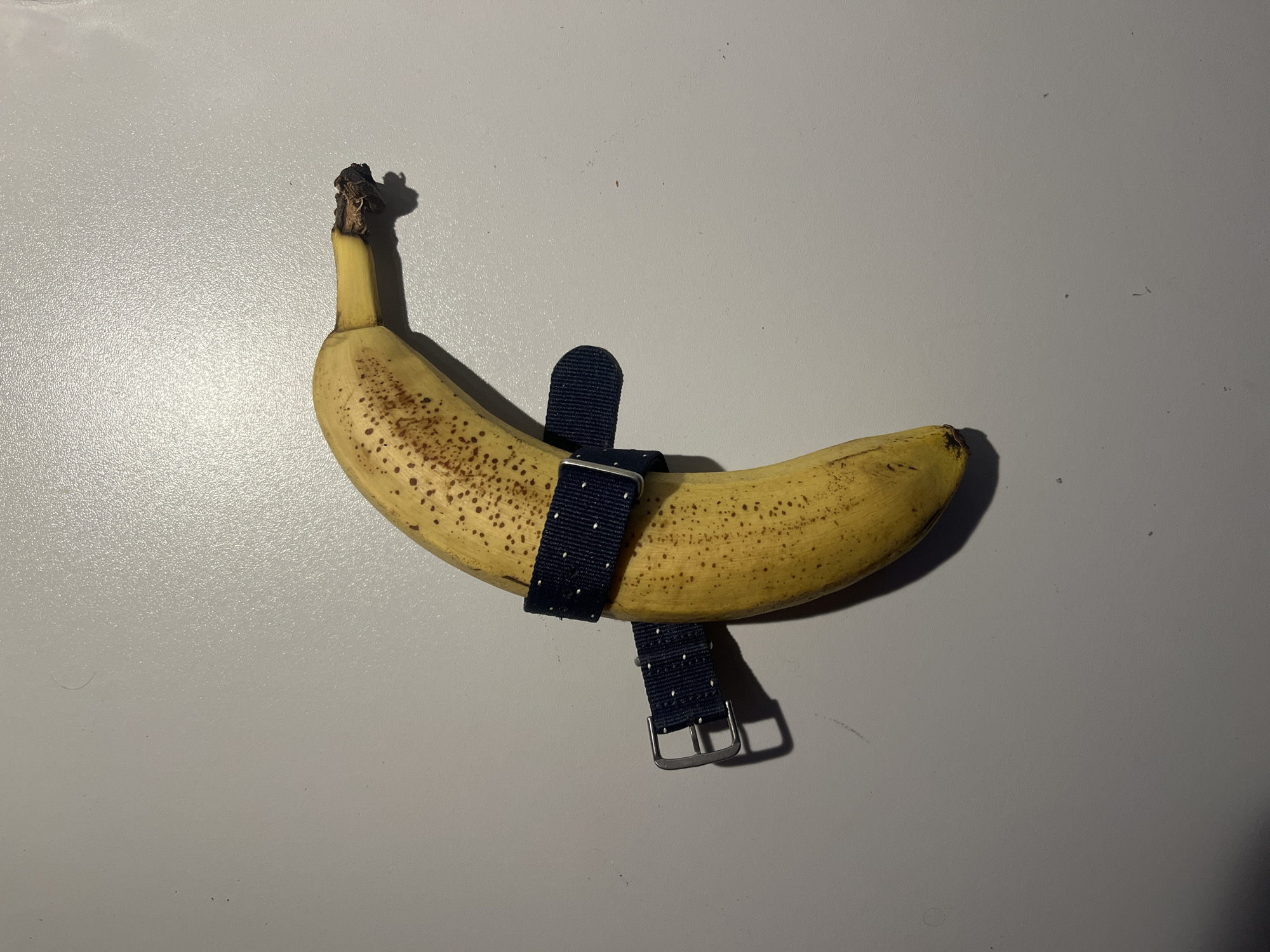 A single, ripe banana with brown spots is positioned diagonally on a plain, light gray surface. A dark blue fabric watch strap with white polka dots is wrapped around the banana, with the buckle and a portion of the strap visible on the left side. The lighting casts shadows to the right of the banana. The scene is indoors, on a textured surface. There is no indication of a city location.