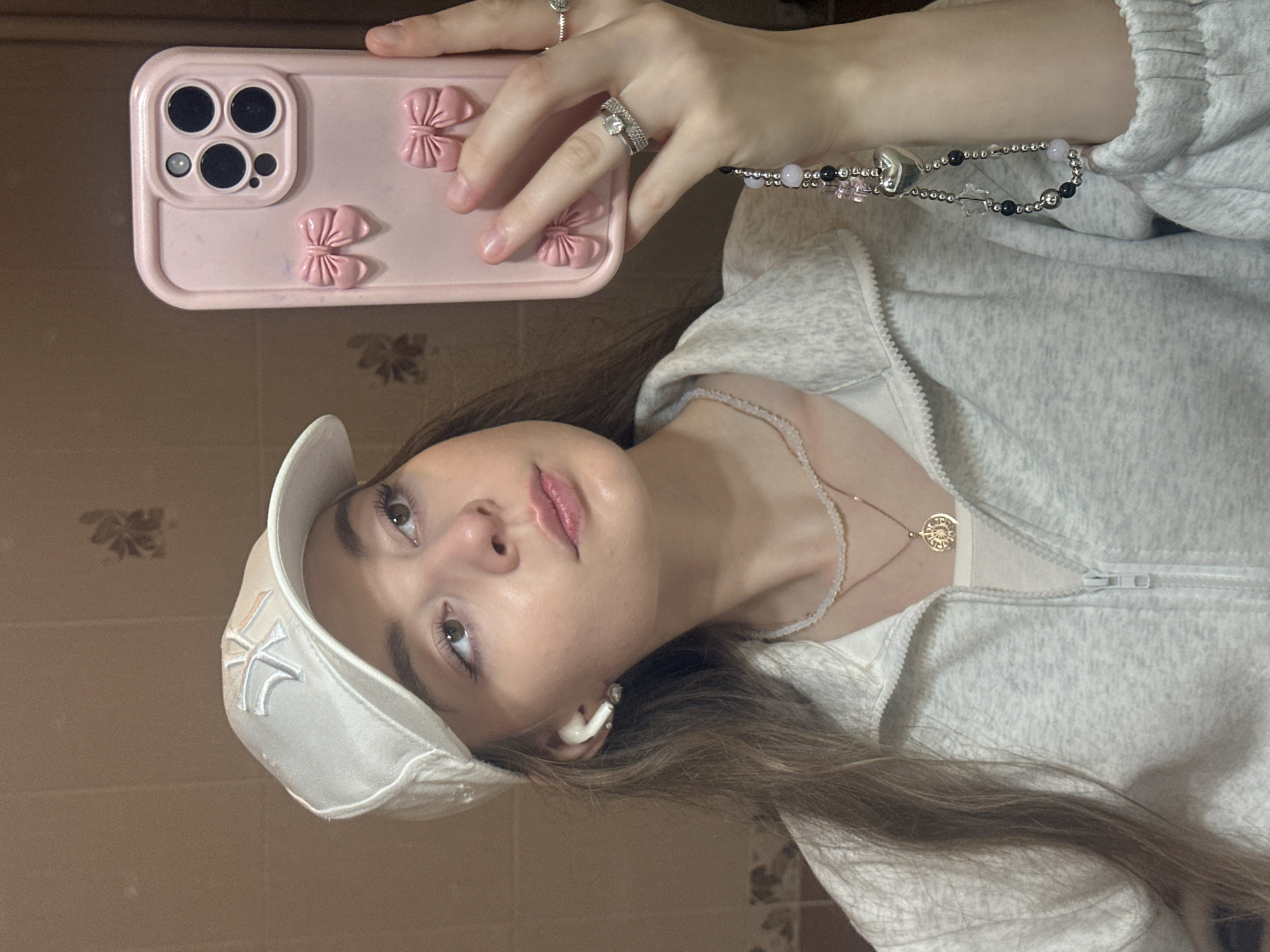 The image shows a young woman taking a selfie in what appears to be a bathroom. She is wearing a white baseball cap with a "NY" logo, a light gray zip-up jacket over a cream-colored t-shirt, and two delicate necklaces. She has white wireless earbuds in her ears and is wearing rings on her fingers. She is holding up a pink iPhone with decorative bows on the case. The background is a plain tiled wall. There are no identifiable landmarks or features that would place this scene specifically in Kazan, Russia.