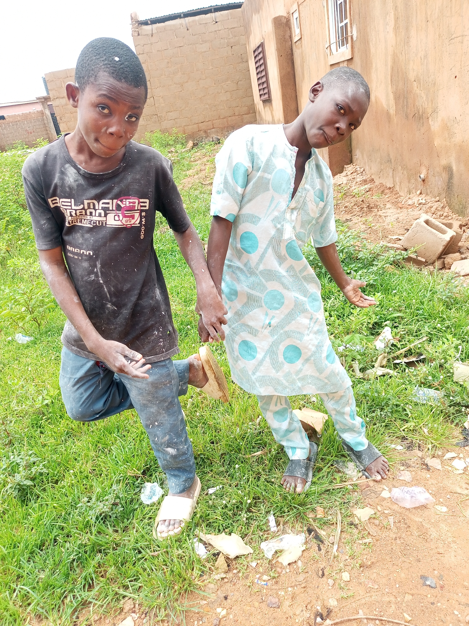 Two young boys stand outdoors, appearing to be in an urban environment in Tempure, Nigeria. The boy on the left wears a black t-shirt and dark pants, with white specks, possibly paint, scattered across his clothing and visible on his face and hair. He has one leg bent, and his right foot is bare, with a sandal placed behind it. The boy on the right is dressed in a light-colored, patterned tunic and matching pants. He is holding the left boy's hand. Both boys are looking towards the camera with somewhat serious expressions. They are standing on a grassy area next to a dirt path. In the background, there is a building constructed with light-colored bricks and a rough, tan-colored wall with windows. Various pieces of debris, including bricks and plastic, are scattered around the ground.