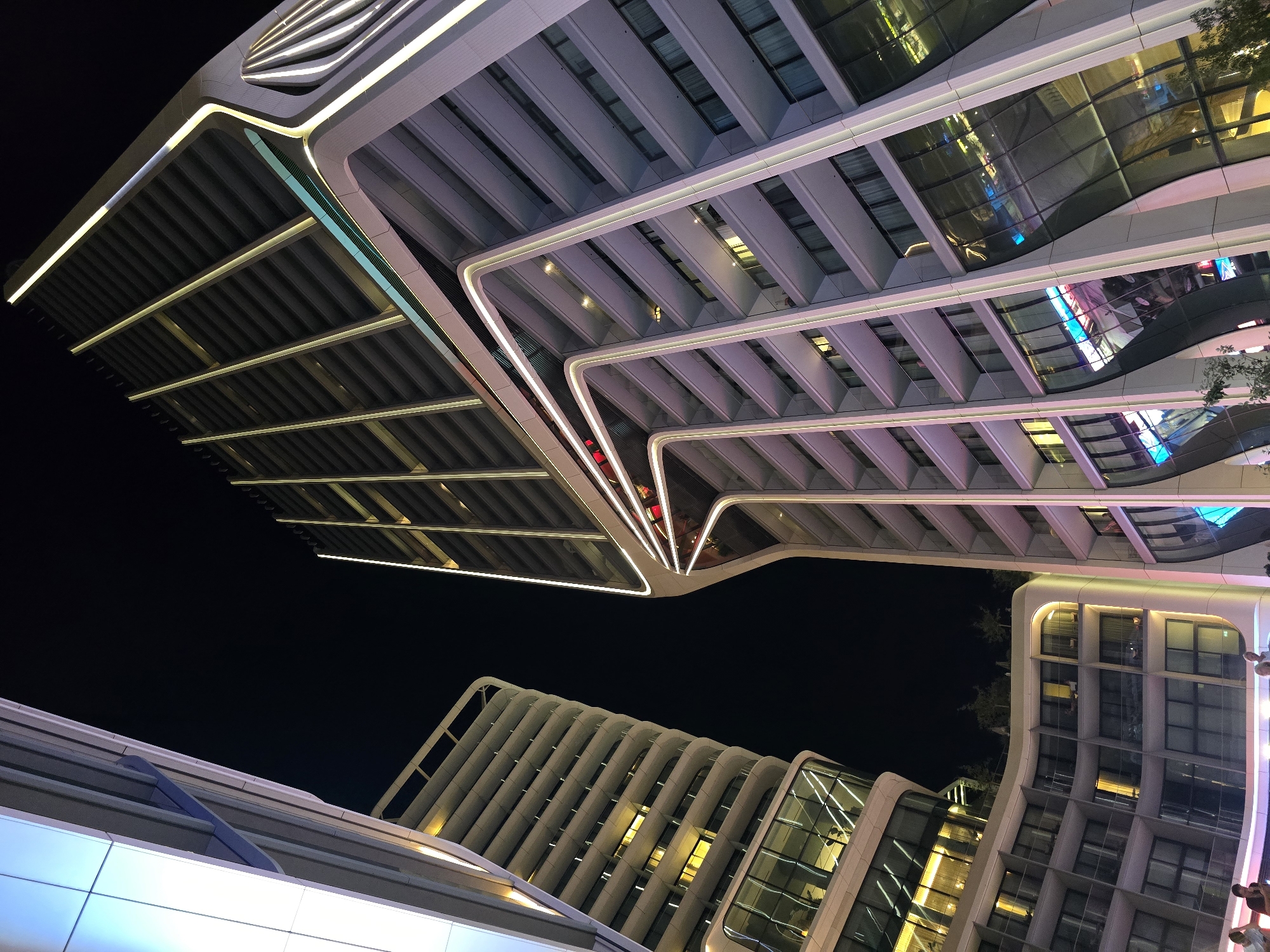 The image captures a nighttime view of the modern architecture of the Portomaso Marina in Saint Julian's, Malta. Two prominent buildings are visible, characterized by their sleek, curved designs and extensive use of lighting. The building on the right is a tall, slender skyscraper with a facade adorned with vertical lines of light, creating a dynamic, glowing effect against the dark night sky. Its lower sections feature large, curved glass windows that reflect the surrounding lights. To the left, another building presents a more rounded, stepped structure, also illuminated with warm, yellow lights in its windows and along its exterior curves. The sky is dark and cloudless. In the foreground, a few individuals are present, silhouetted against the bright lights of the buildings, suggesting they are pedestrians or residents. The overall scene conveys a sense of urban sophistication and modern design.