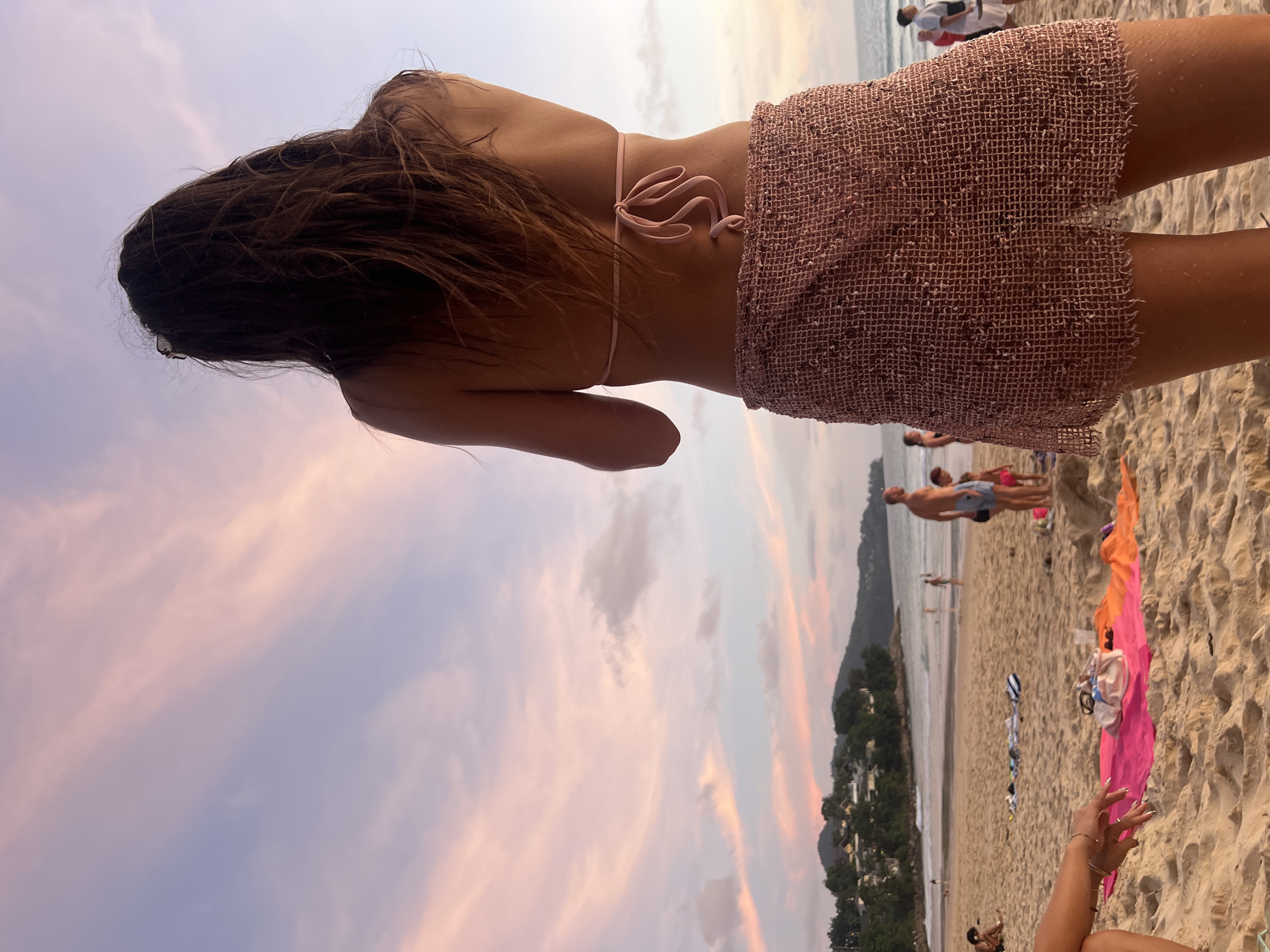 A young woman with long, dark hair stands on a sandy beach in Choeng Thale, Thailand, facing away from the camera and looking out at the ocean. She is wearing a pink bikini top and a short, textured pink skirt. The sky above is filled with soft, pastel clouds, indicating either sunrise or sunset. In the background, there are other beachgoers, some walking along the shore and others relaxing on the sand. Waves are gently rolling onto the beach, and the coastline features a line of trees and hills. The overall scene depicts a serene beach environment at dusk or dawn.