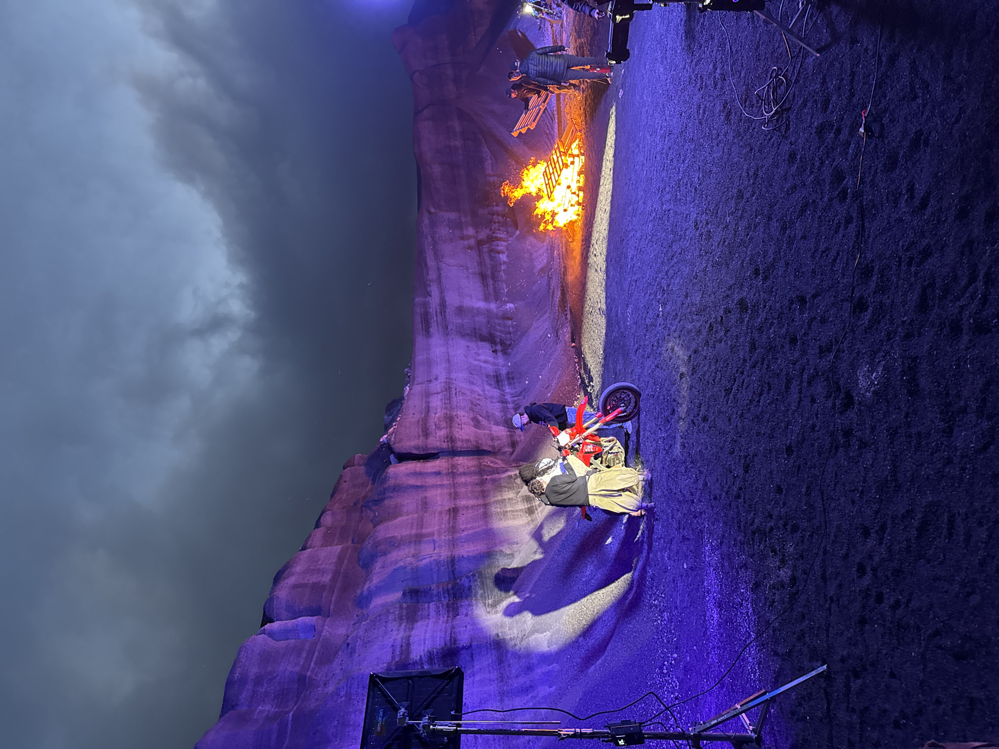The image depicts an outdoor scene, likely at night or dusk, possibly in the vicinity of Teguise, Spain, although this cannot be definitively verified from the image alone. A large rock formation, illuminated with purple light, dominates the left side of the frame. Adjacent to the rock formation, a fire burns brightly. Two or three individuals are positioned near a red dirt bike. One person seems to be near the front wheel, perhaps working on the bike. The ground is dark and covered with what appears to be gravel. In the foreground, lighting equipment is visible, indicating a film or photo shoot. The sky above is overcast.