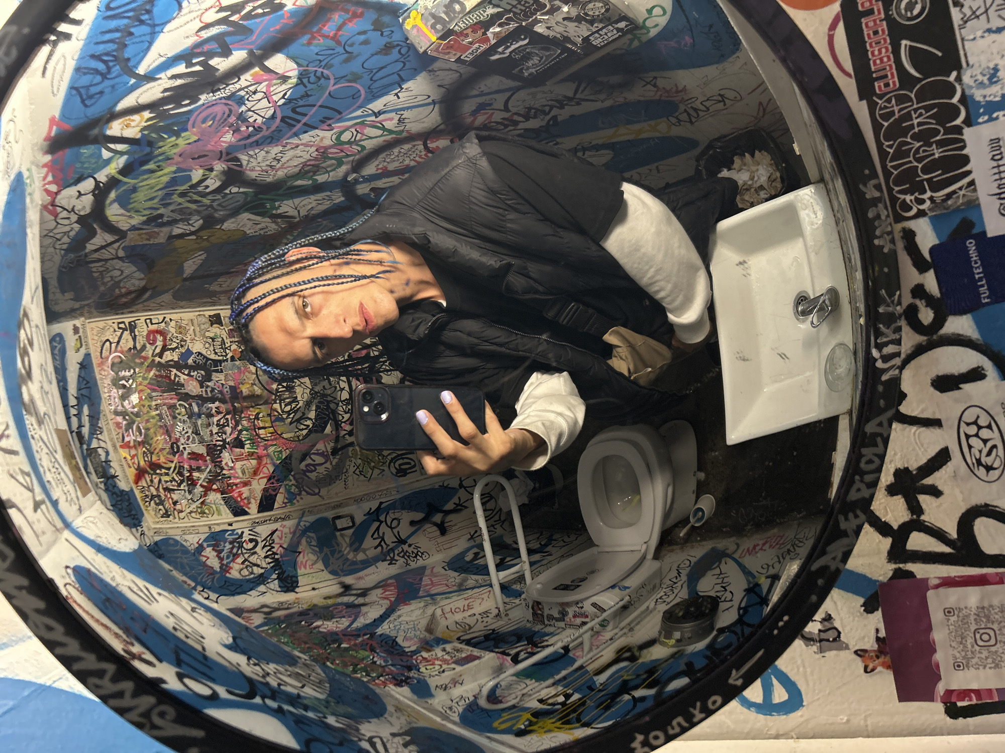 The image is a selfie taken in a toilet stall, likely in a public venue, as suggested by the extensive graffiti covering the walls and ceiling. The person in the selfie has light skin, dark hair styled in braids with blue highlights, and is wearing a black puffer jacket over a white long-sleeved shirt. They are holding a black smartphone, presumably to take the picture.
The toilet stall contains a toilet with a child seat, a sink, and a trash can overflowing with paper towels. Stickers and graffiti cover every surface, including the toilet and the sink. The graffiti appears to be in a mix of blue, black, pink, and other colors. Stickers and writing are in several languages.
The presence of "ALE PIOLA" and "CLUBSOCALP" stickers may suggest a location in the city of Buenos Aires, Argentina. These are likely associated with local cultural events or businesses.