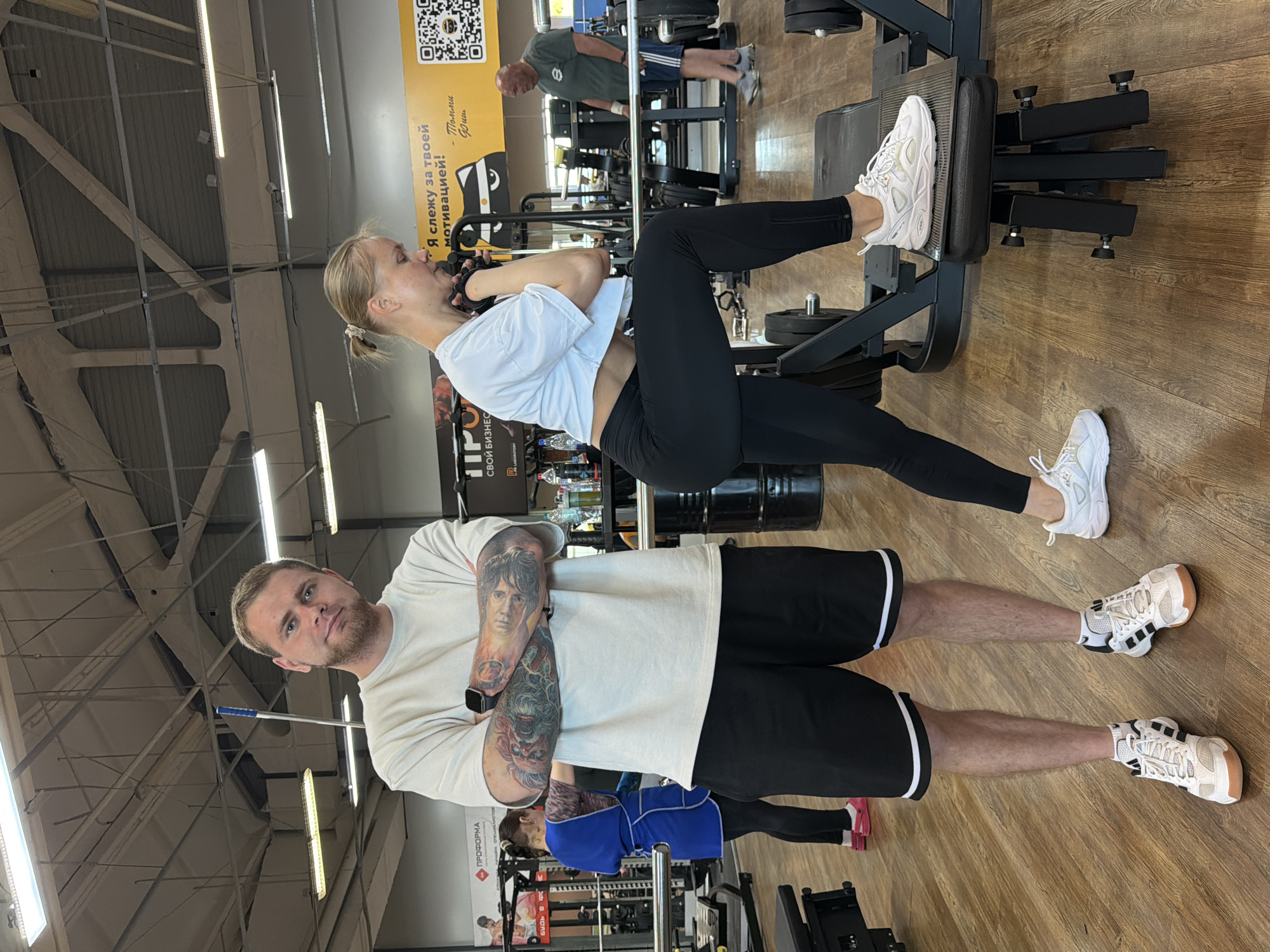 The image depicts an indoor gym setting, likely in Chaikovsky, Russia, given the presence of Russian text on a banner in the background. In the foreground, a man with tattoos on his arms stands with his arms crossed, facing the camera. He is wearing a light-colored t-shirt and black shorts. To his right, a woman is performing a strength exercise, with one foot elevated on a piece of gym equipment. She is wearing a white crop top and black leggings. In the background, other individuals are visible, engaged in their workouts or spectating. The gym environment includes various weightlifting machines, benches, and weight plates, characteristic of a commercial fitness facility. The lighting suggests it is daytime, with artificial lights illuminating the space. The overall atmosphere appears to be one of active fitness pursuits.