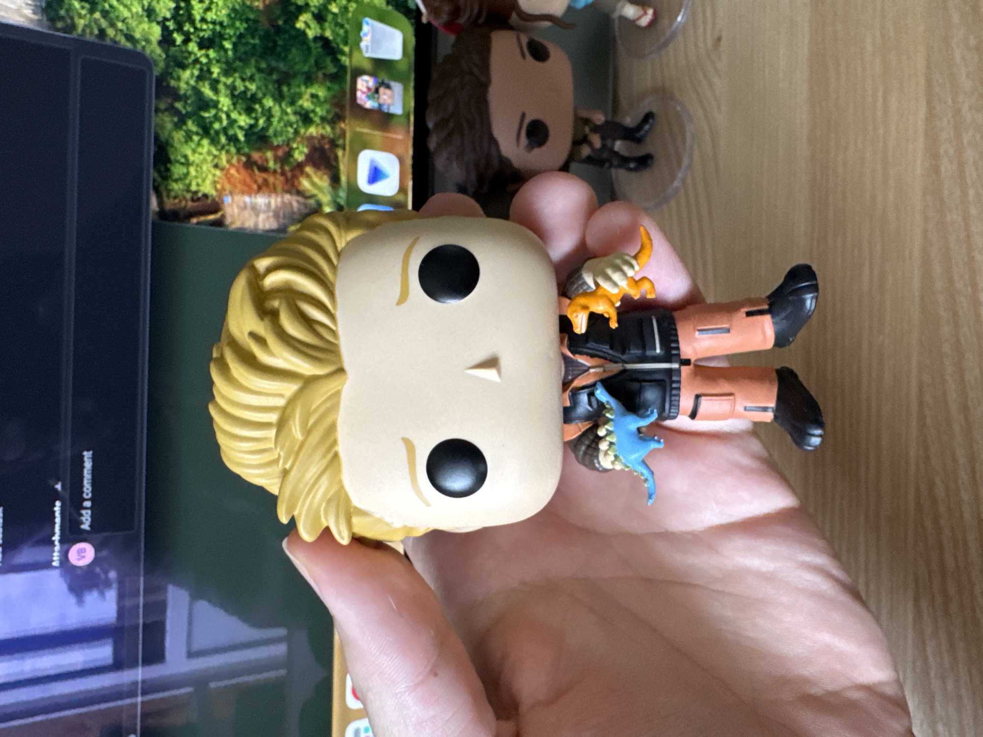 A close-up shot focuses on a hand holding a Funko Pop figure of Chris Pratt as Owen Grady from Jurassic World. The figure is depicted with its signature blonde hair, wearing an orange and black outfit, and holding two small, colorful dinosaur figures: a blue one in its left hand and an orange one in its right. In the background, other Funko Pop figures are visible, including a female figure with dark hair and another figure with brown hair. A monitor screen displays what appears to be a website interface, with elements like "Attachment" and "Add a comment" visible, suggesting an online context. The scene is set against a wooden surface. The image does not provide any information regarding the city of Bila Tserkva, Ukraine.